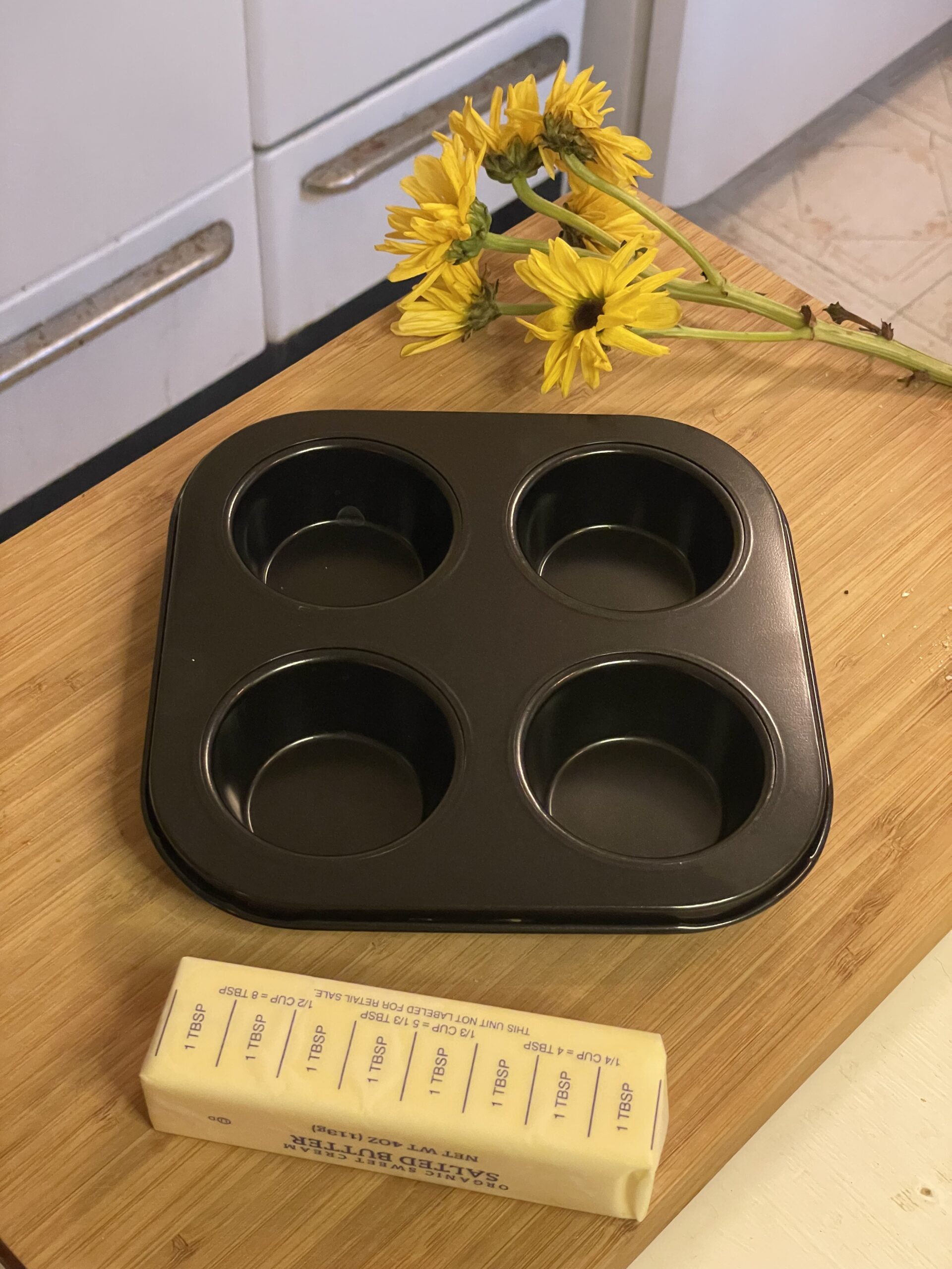 A black nonstick muffin tin sits beside a stick of salted butter and a cluster of yellow daisies on a wooden surface, evoking a cozy, vintage kitchen scene.