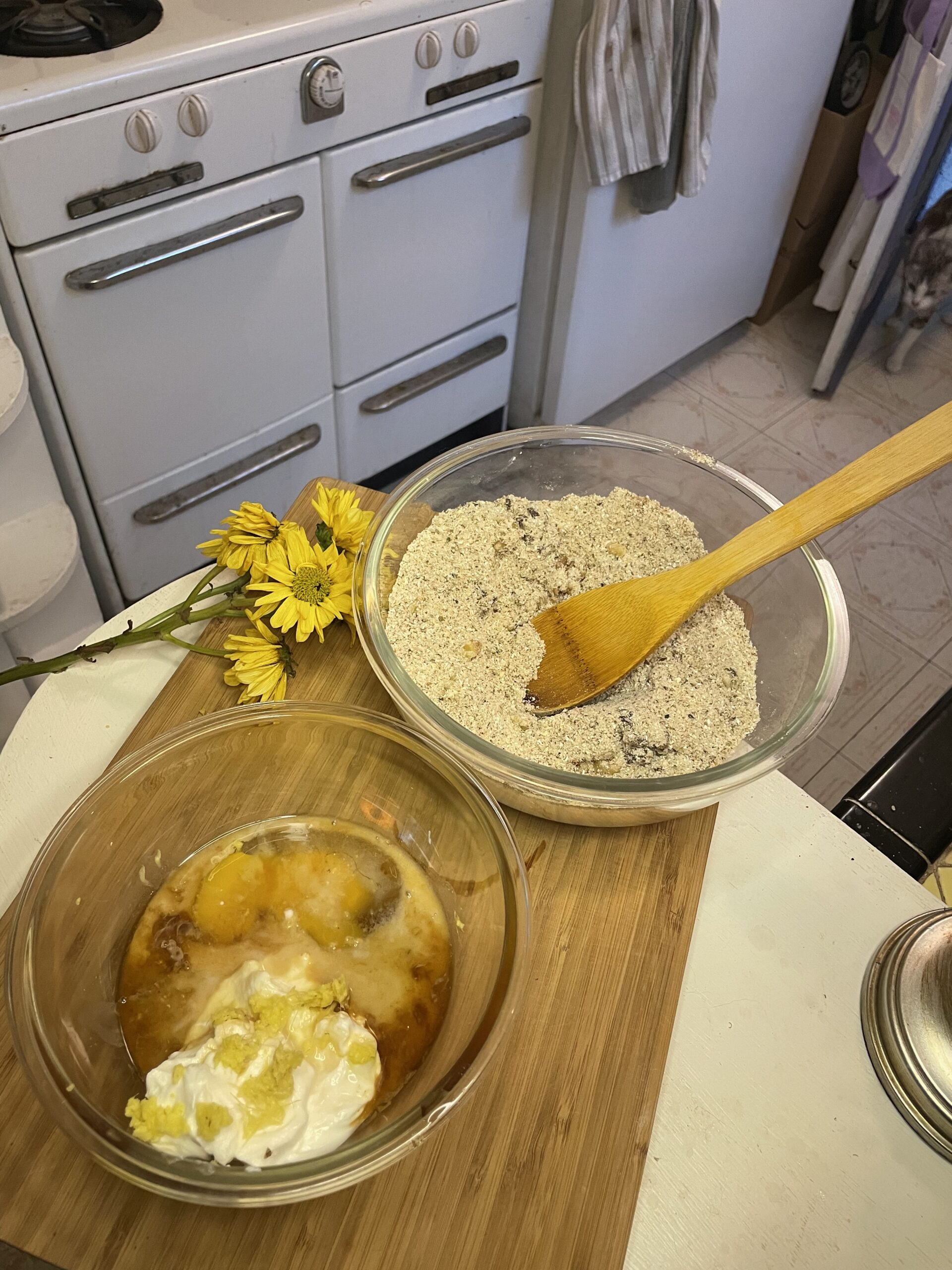 A glass bowl filled with a spiced dry muffin mix sits on a butcher block counter next to a small bunch of yellow daisies, ready for wet ingredients to be added.