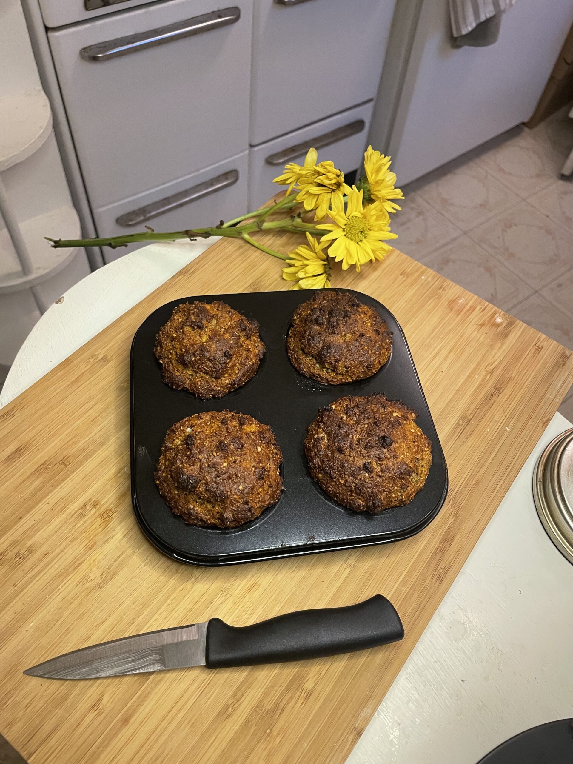 Freshly baked muffins with a rustic golden crust resting in a black muffin tin, surrounded by a yellow daisy bouquet and a paring knife on a wooden cutting board.