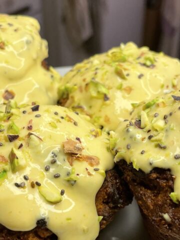 Close-up of four golden-brown muffins topped with a vibrant ginger-turmeric glaze and crushed pistachios, served on a white plate in a warmly lit kitchen.