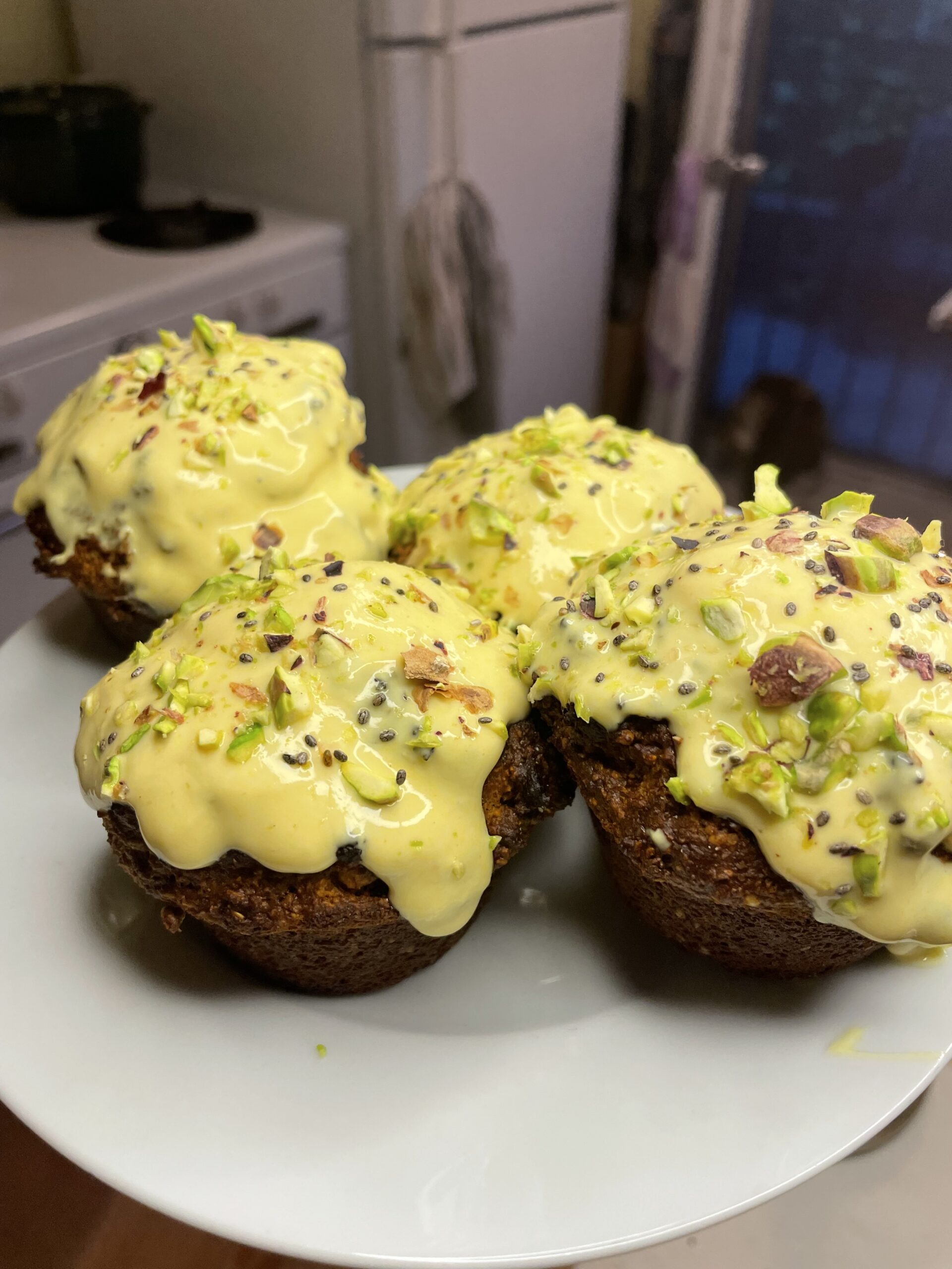Close-up of four golden-brown muffins topped with a vibrant ginger-turmeric glaze and crushed pistachios, served on a white plate in a warmly lit kitchen.