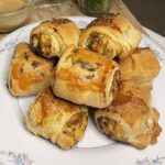 Golden-brown herb-crusted chicken and apple sausage rolls stacked on a floral-trimmed white plate, showcasing a flaky puff pastry exterior brushed with egg wash and topped with fresh herbs.