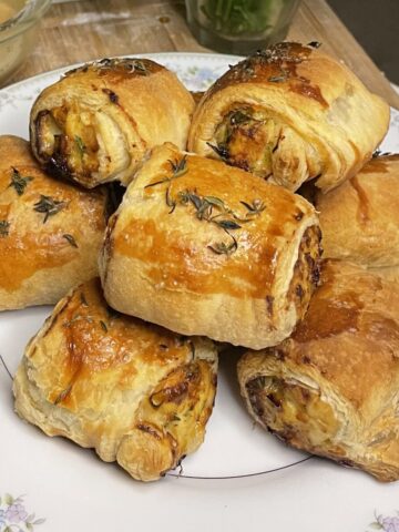 Golden-brown herb-crusted chicken and apple sausage rolls stacked on a floral-trimmed white plate, showcasing a flaky puff pastry exterior brushed with egg wash and topped with fresh herbs.