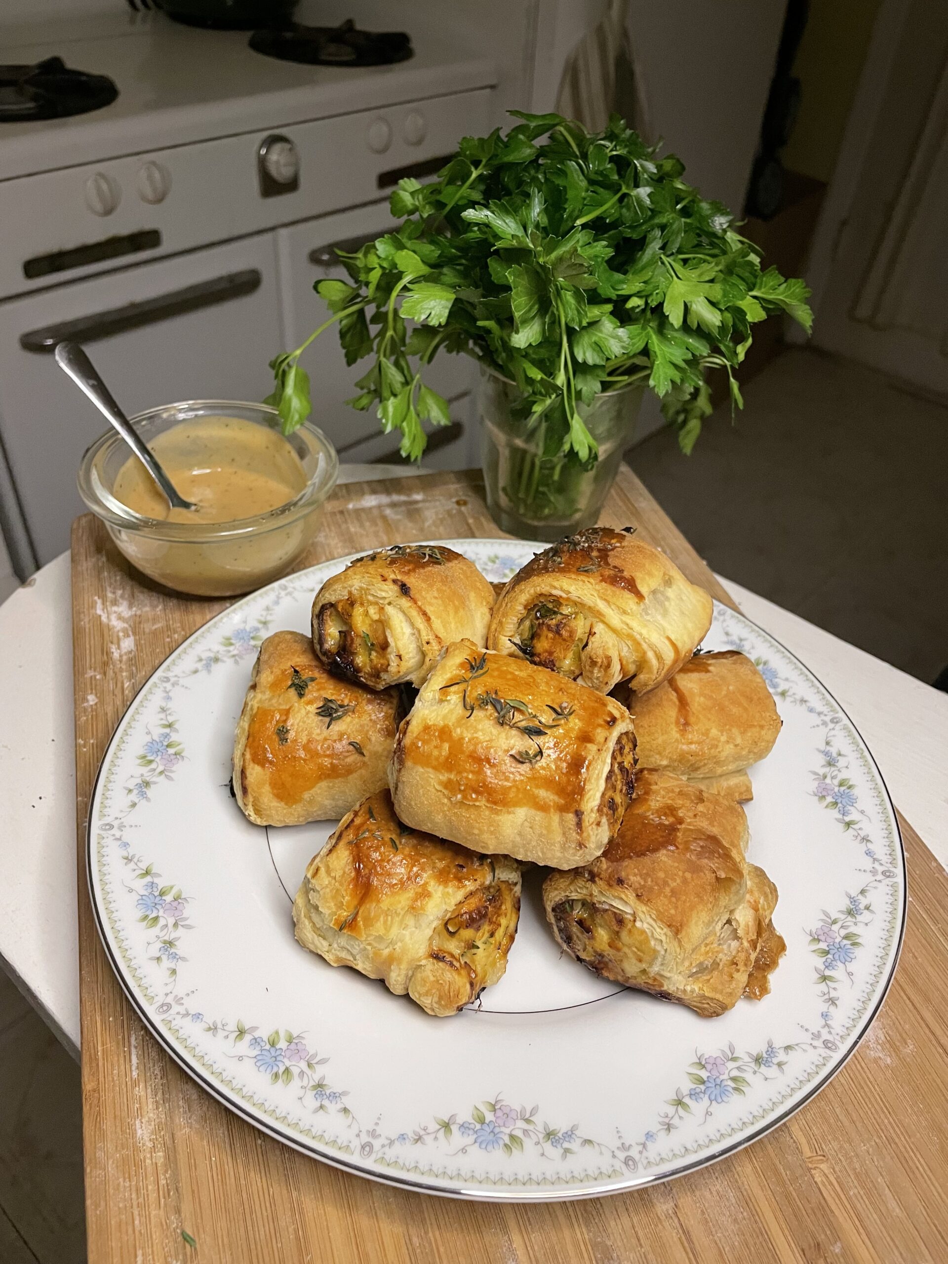 Golden-brown herb-crusted chicken and apple sausage rolls stacked on a floral-trimmed white plate, showcasing a flaky puff pastry exterior brushed with egg wash and topped with fresh herbs.