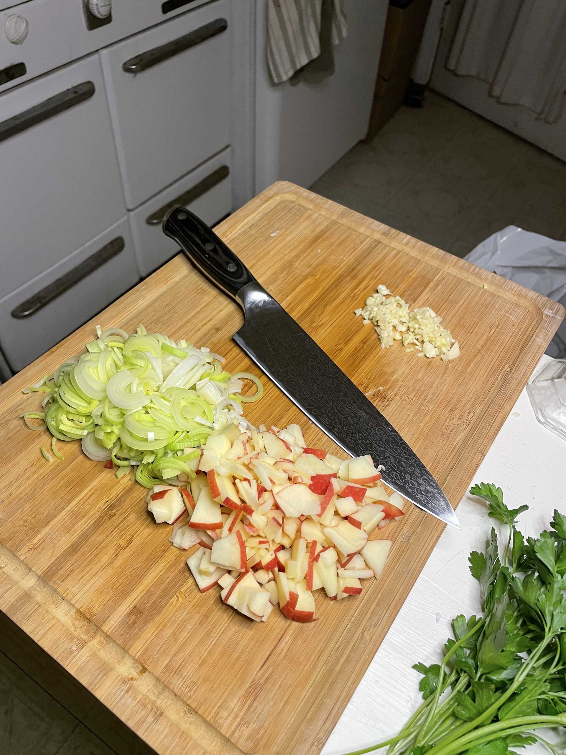 A wooden cutting board with finely chopped leeks, diced red-skinned apples, and minced garlic beside a chef’s knife, set in a cozy kitchen environment with a bunch of parsley in the corner.