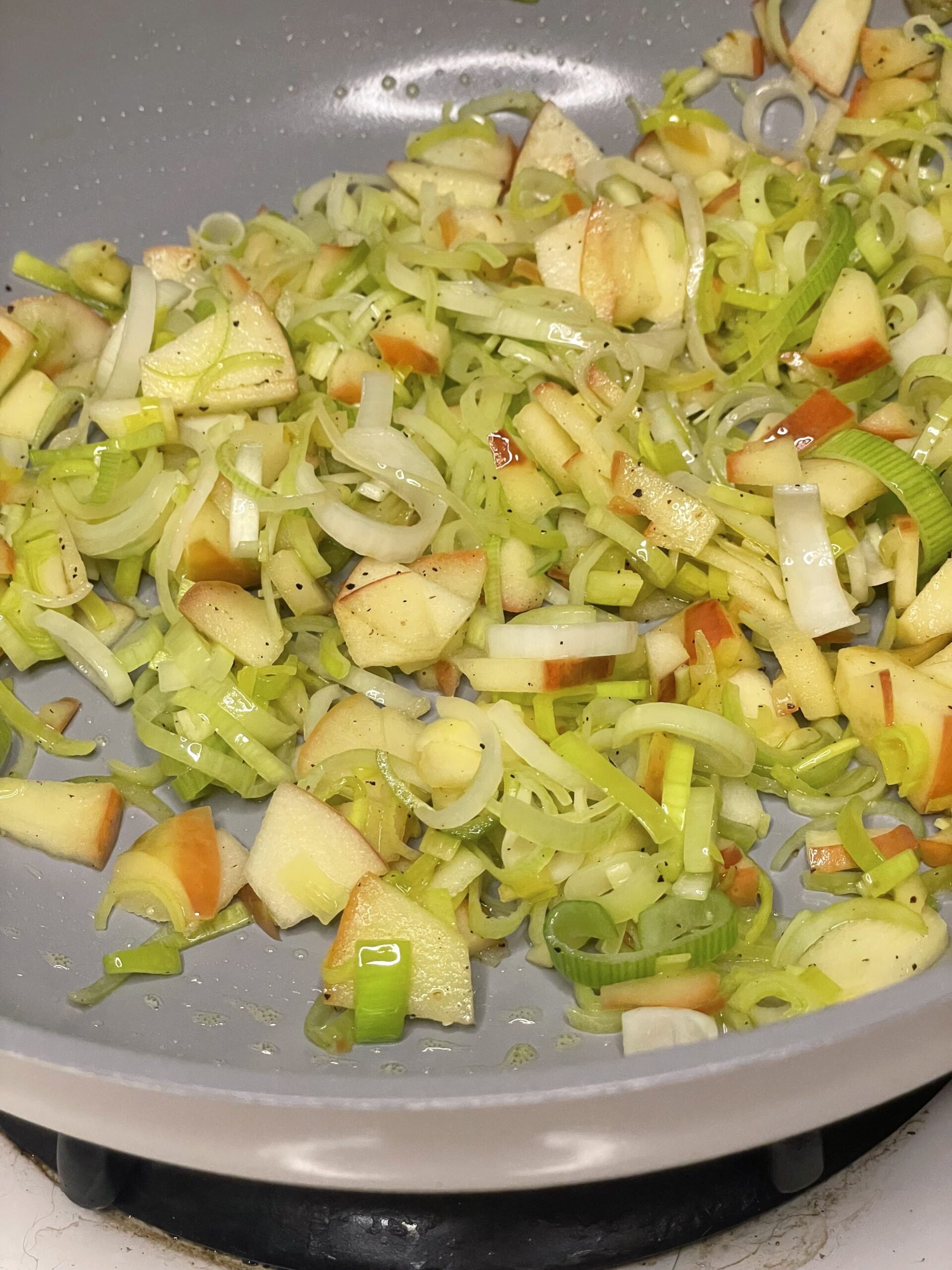 A close-up of sliced leeks and diced apples being sautéed in a non-stick skillet, lightly seasoned and glistening with oil as they begin to soften and caramelize.