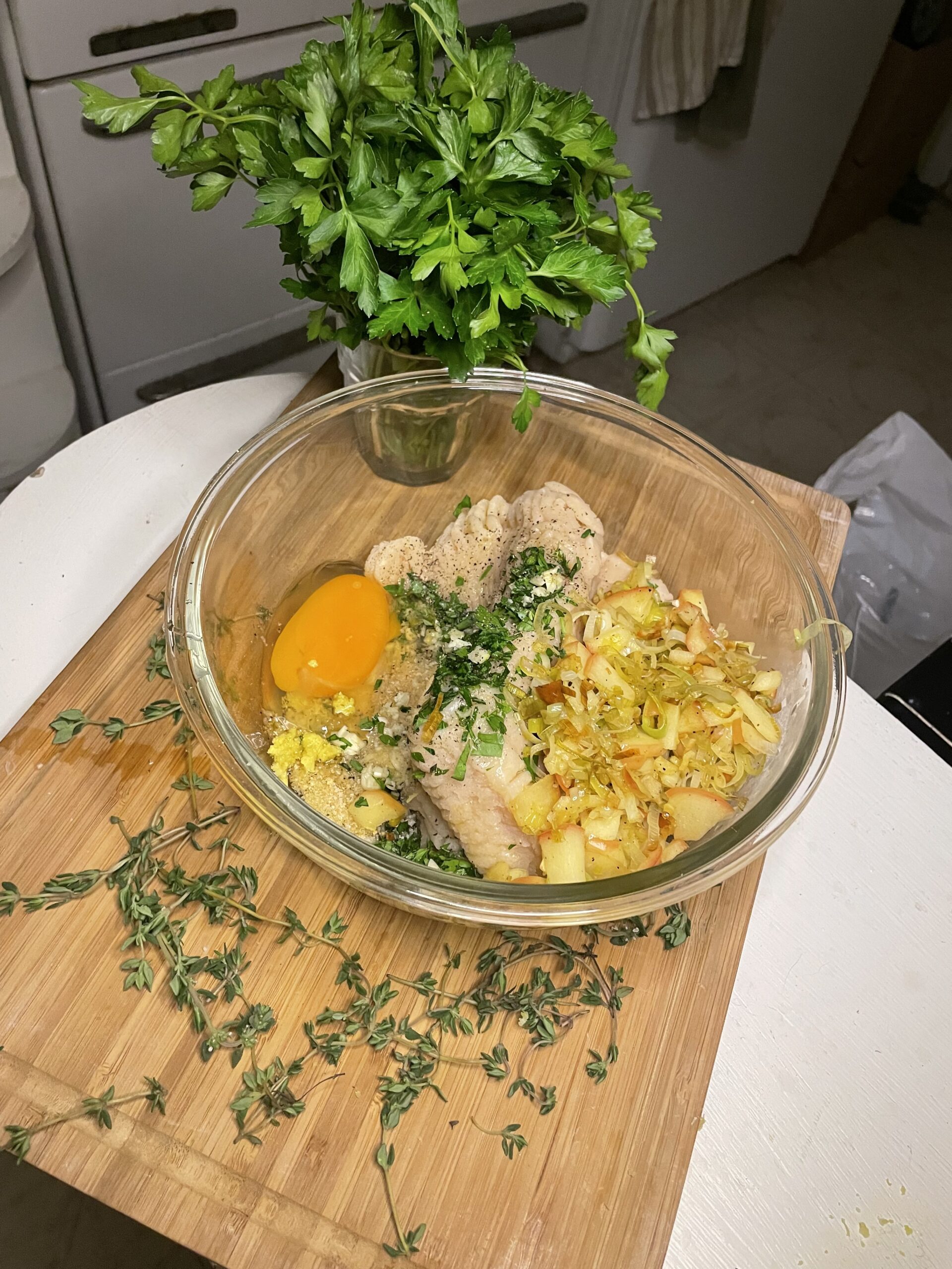 A glass mixing bowl containing ground chicken, an egg yolk, sautéed leeks and apples, and chopped fresh herbs like thyme and parsley, ready to be combined into a flavorful filling.