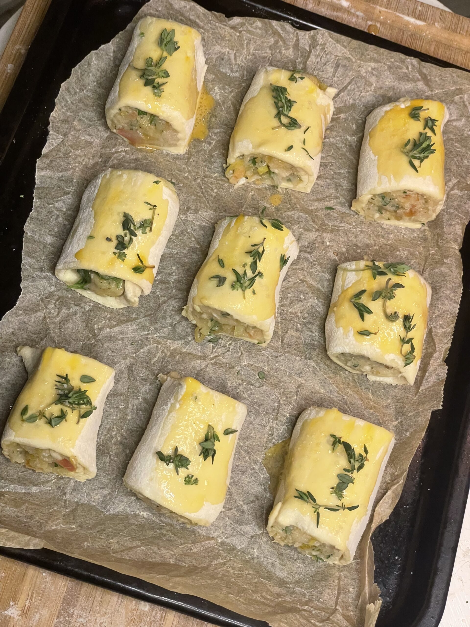 Unbaked puff pastry rolls filled with chicken-apple mixture arranged neatly on parchment paper, each brushed with egg wash and sprinkled with fresh herbs, ready to go into the oven.