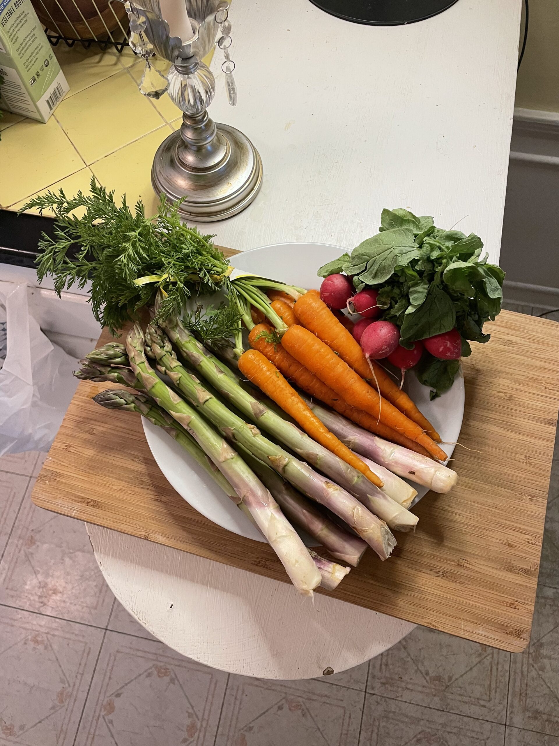 Plate piled high with fresh spring vegetables including carrots with greens, radishes, and thick asparagus spears, sitting atop a wooden cutting board on a kitchen counter.