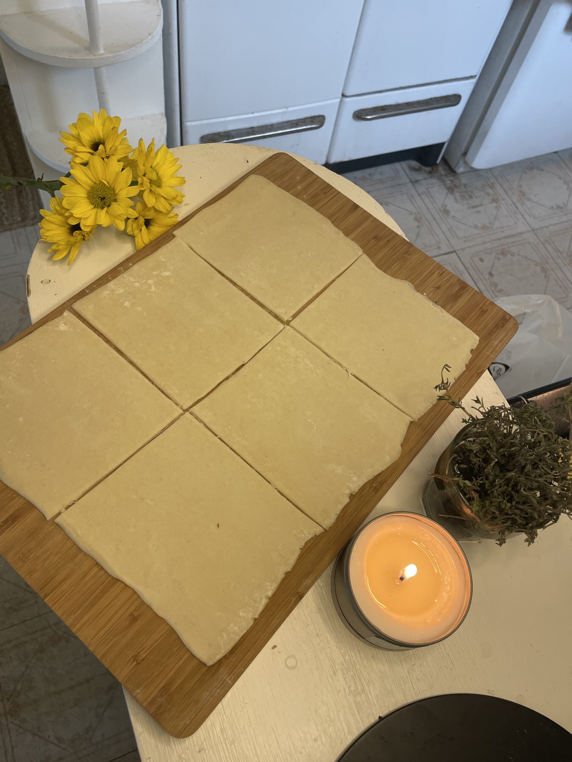 Unbaked puff pastry dough cut into equal square sections on a wooden board, next to a bunch of yellow flowers and a lit candle, ready for assembling.