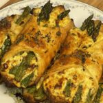 Golden brown puff pastry bundles filled with roasted asparagus and herbed ricotta cheese, served on a floral porcelain plate atop a wooden board.