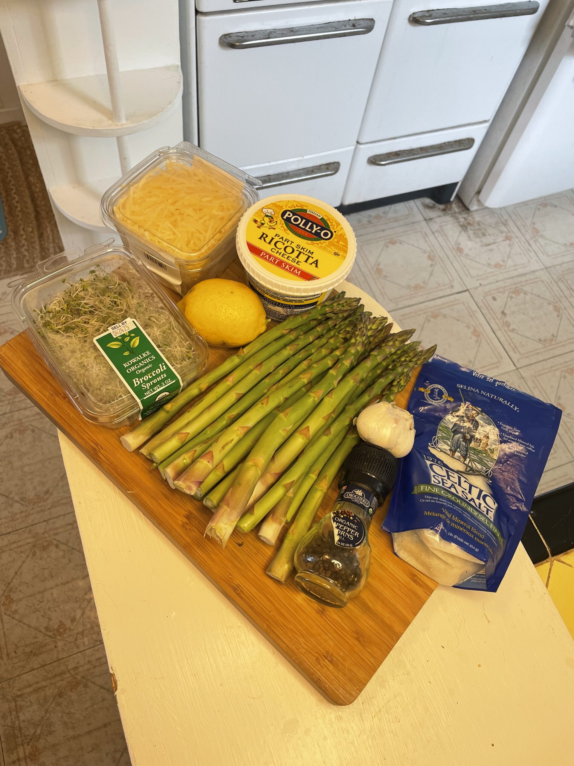 Fresh ingredients for asparagus ricotta bundles arranged on a wooden board, including a bunch of asparagus, part-skim ricotta, shredded cheese, garlic, lemon, black pepper, sea salt, and broccoli sprouts.