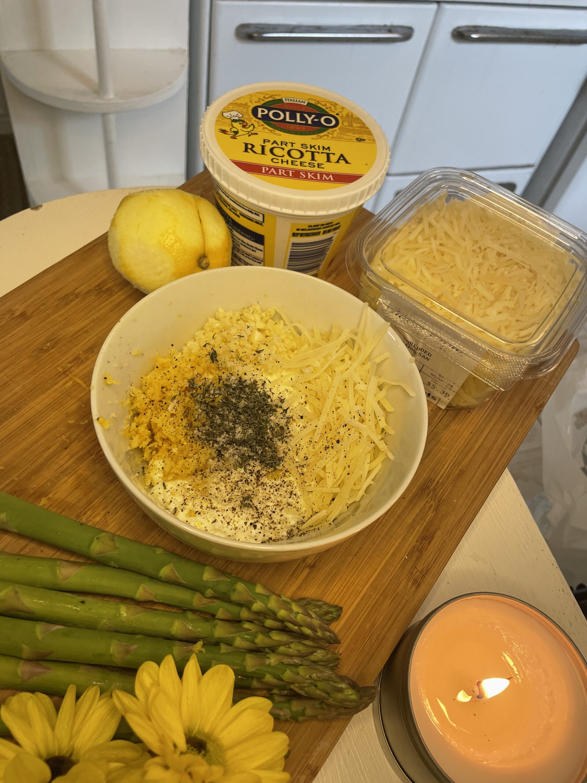 Close-up of a ricotta cheese mixture in a white bowl with shredded cheese, lemon zest, cracked black pepper, and dried herbs, surrounded by asparagus, a lemon, and a lit candle.