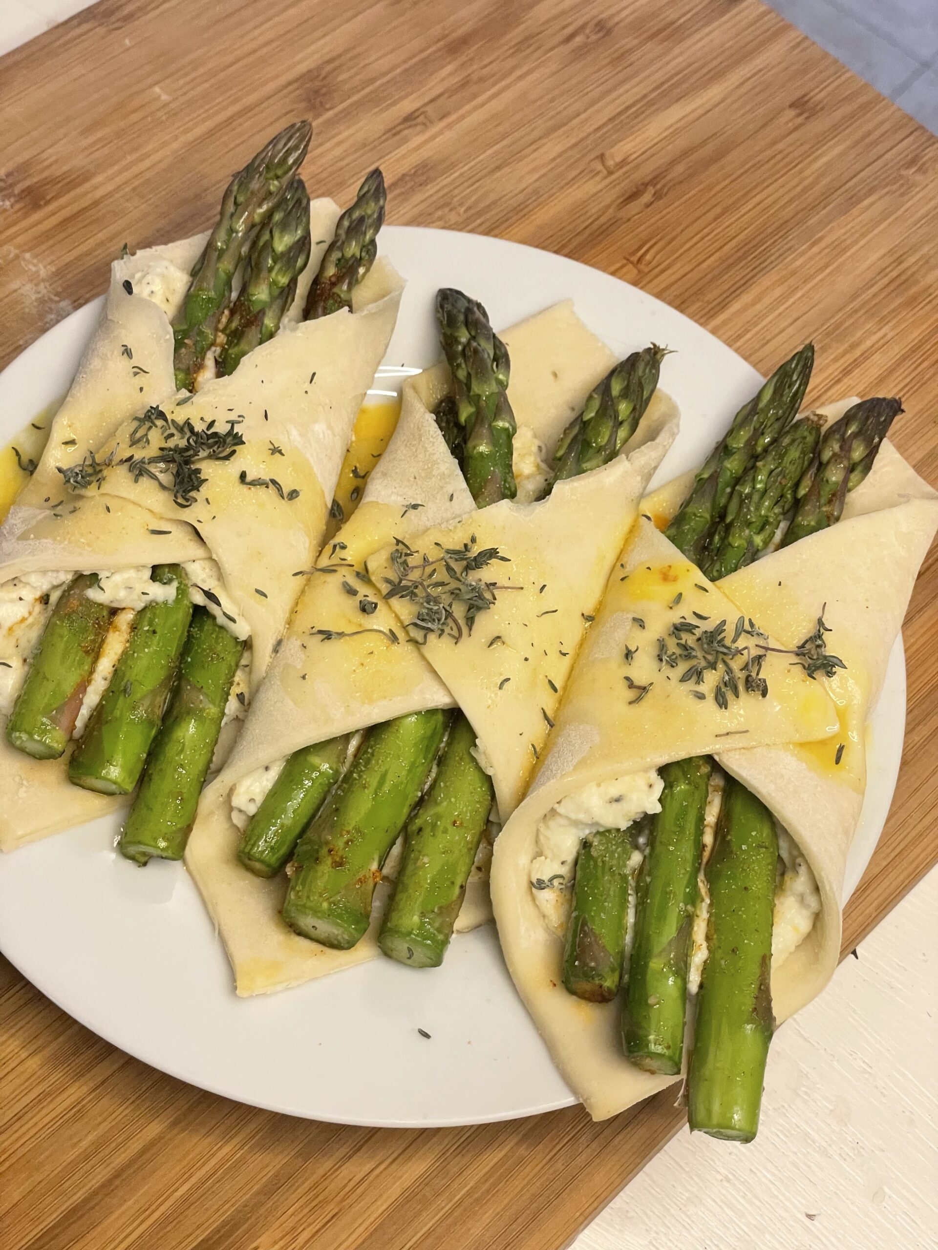 Pre-baked asparagus and ricotta bundles wrapped in pastry dough, brushed with egg wash and sprinkled with herbs, arranged neatly on a white plate.