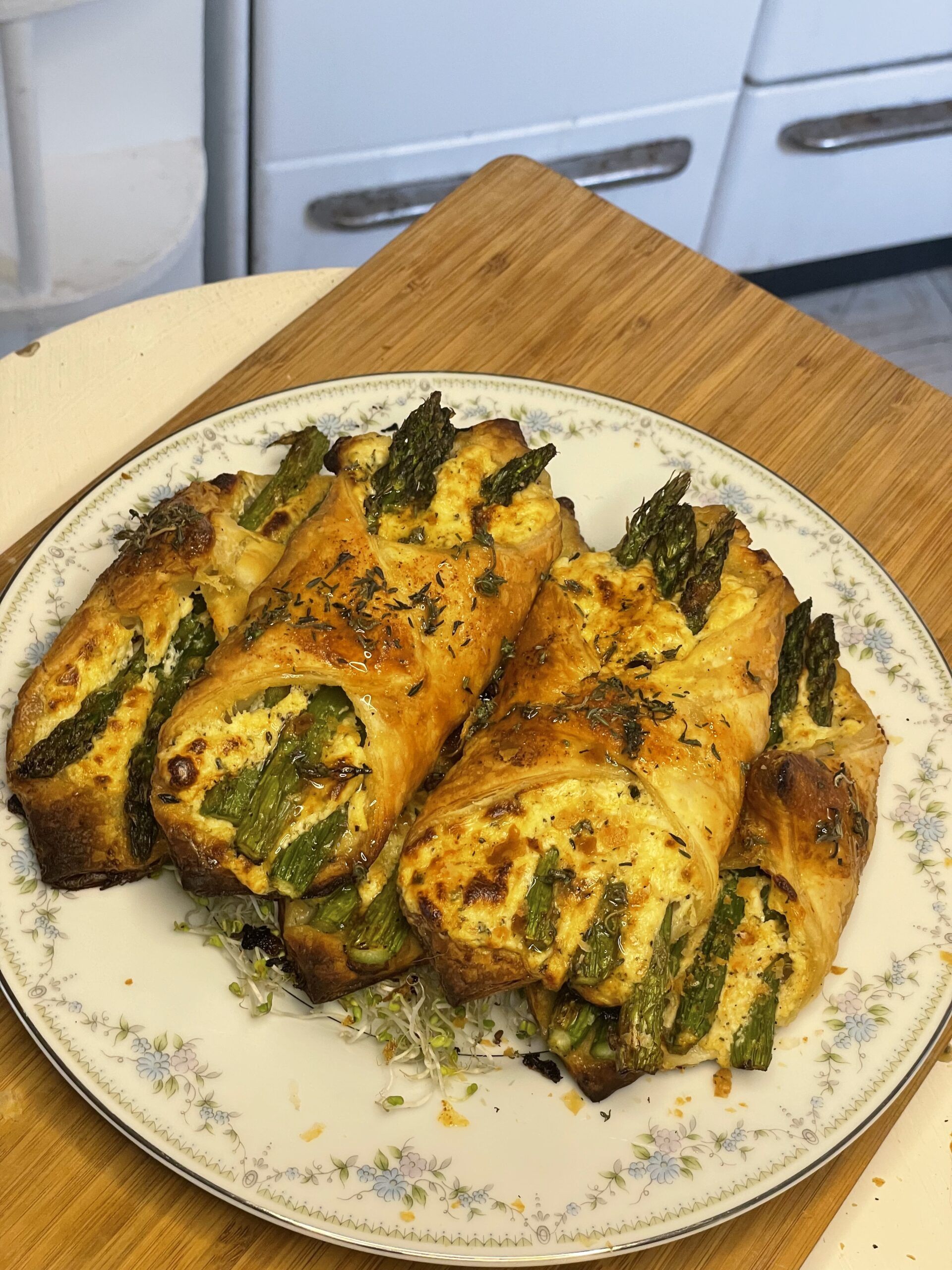 Golden brown puff pastry bundles filled with roasted asparagus and herbed ricotta cheese, served on a floral porcelain plate atop a wooden board.