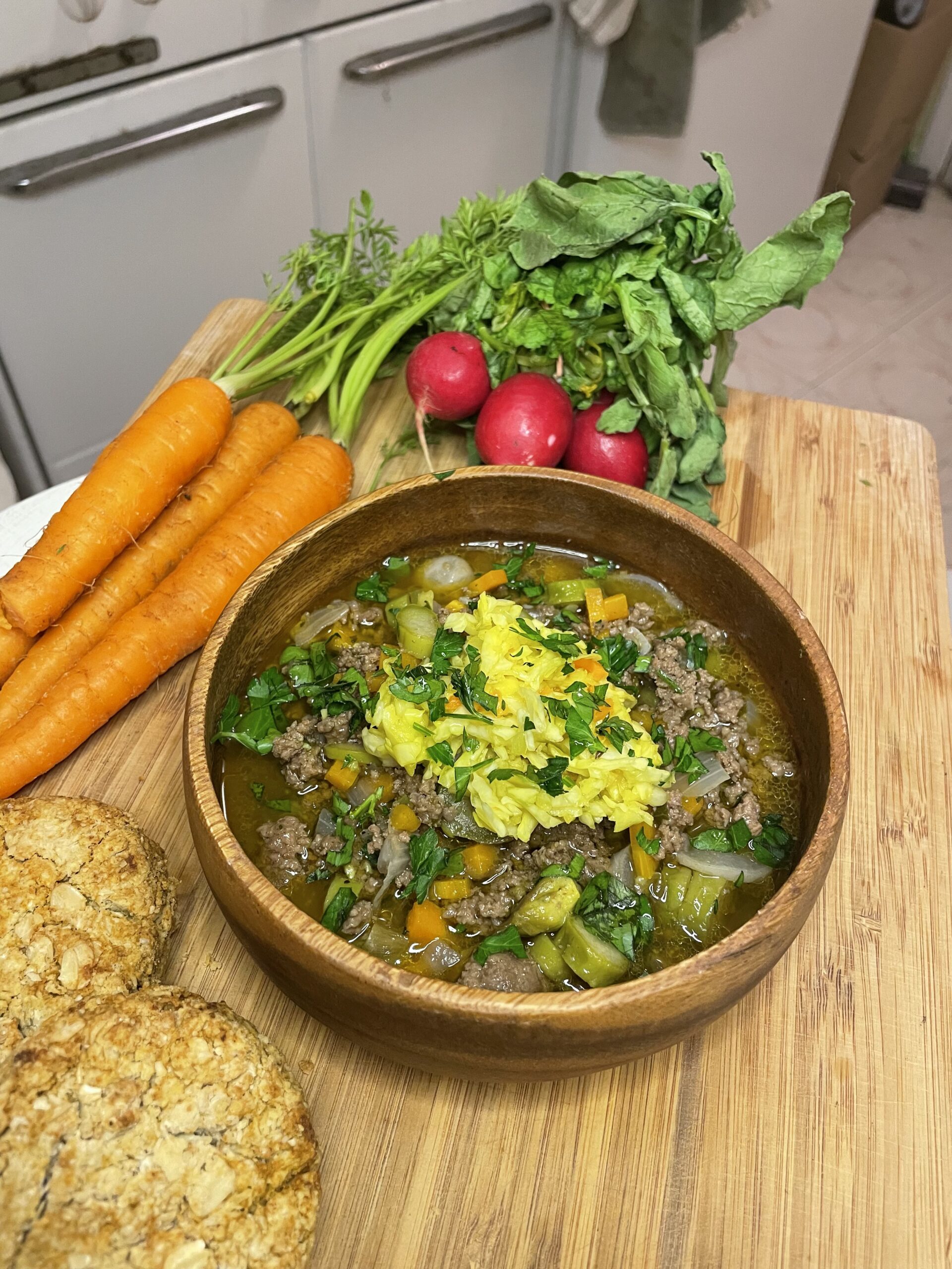 Rustic bowl of springtime beef and vegetable stew with yellow cabbage garnish, placed next to fresh carrots, radishes, and oat biscuits on a wooden cutting board.