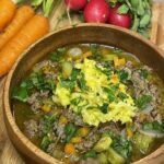 Hearty bowl of springtime beef and vegetable stew topped with shredded yellow cabbage and fresh herbs, surrounded by fresh carrots and radishes on a wooden cutting board.