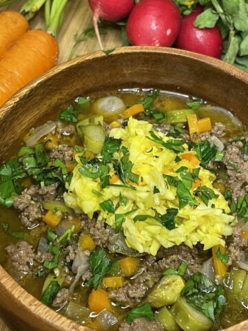 Hearty bowl of springtime beef and vegetable stew topped with shredded yellow cabbage and fresh herbs, surrounded by fresh carrots and radishes on a wooden cutting board.