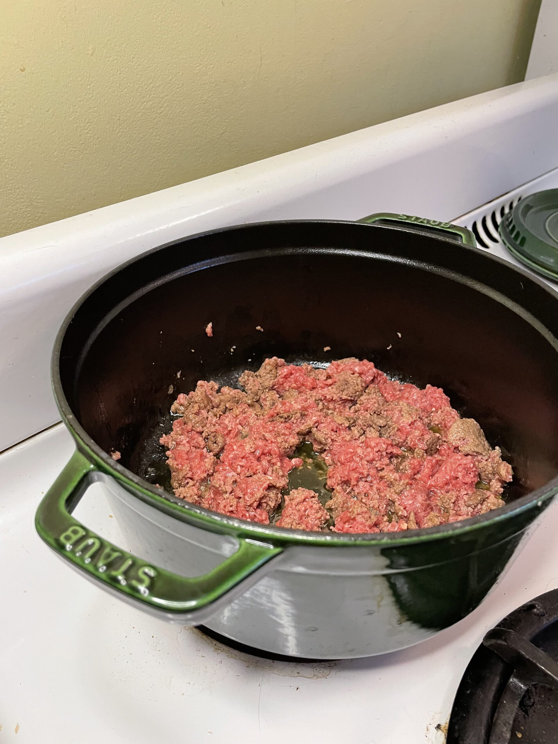 Ground beef sizzling in a green Staub Dutch oven on a white stovetop, partially browned with pink sections still visible.