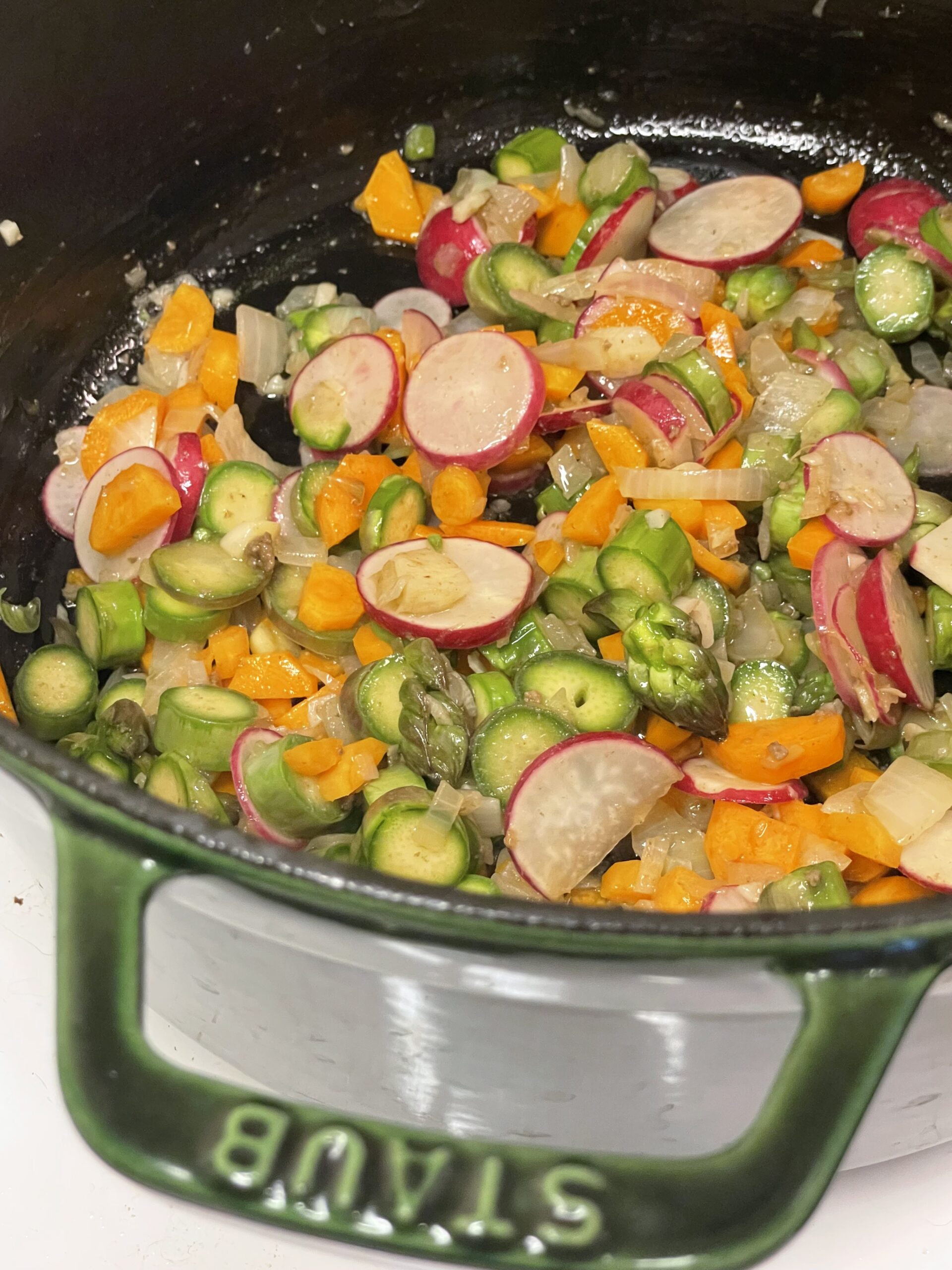 Sautéed chopped vegetables including sliced radishes, carrots, and asparagus in a green Staub Dutch oven, lightly cooked and vibrant.