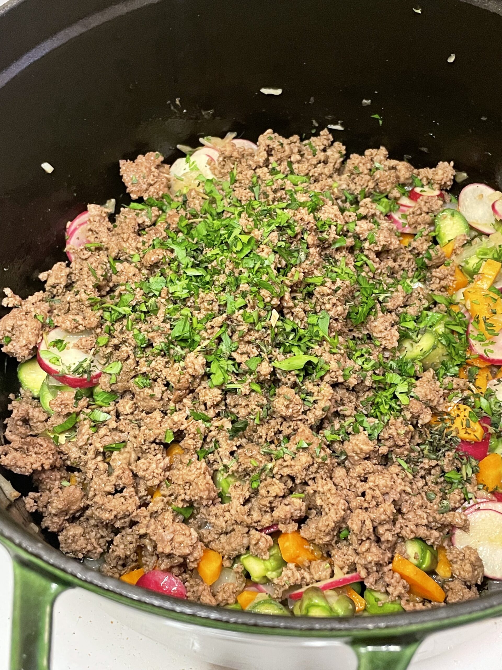 Fully browned ground beef layered over sautéed vegetables in a green Staub Dutch oven, topped with a sprinkle of chopped fresh herbs.