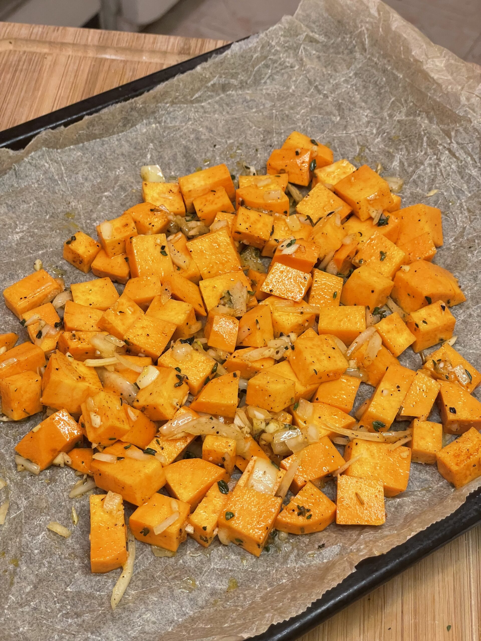 Cubed orange squash and slivered onions seasoned with herbs and spices, spread evenly across a parchment-lined baking sheet for roasting.