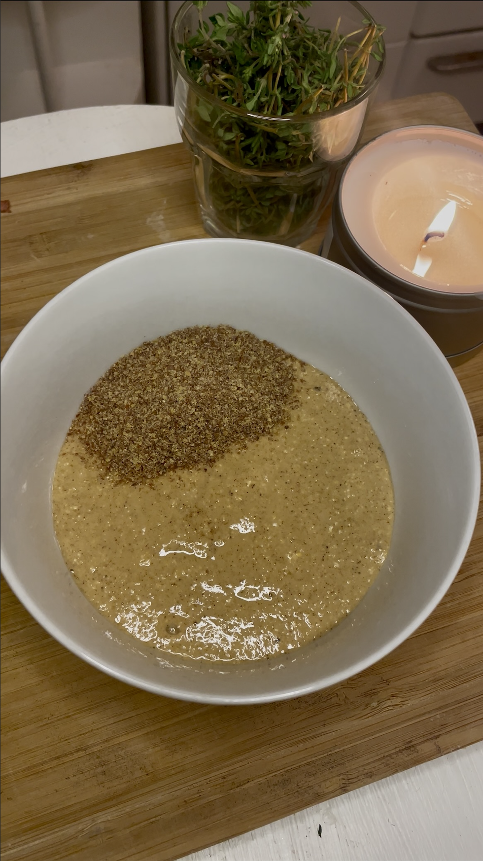 A white bowl on a wooden surface containing a thick golden batter partially topped with ground flaxseed, with fresh herbs and a candle in the blurred background.