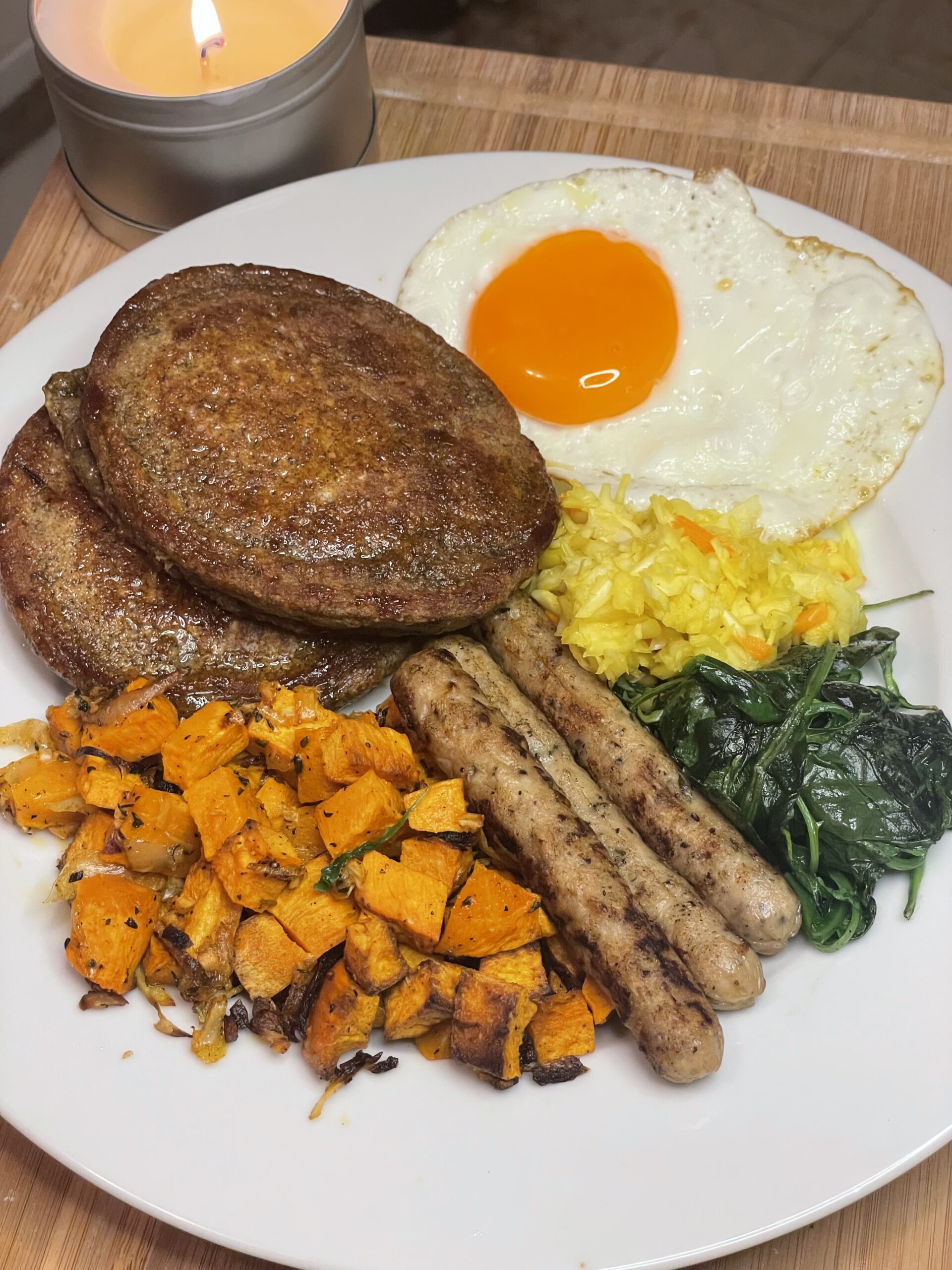 A hearty breakfast plate featuring sweet potato hash, chicken sausages, golden oat pancakes, sautéed spinach, turmeric rice, and a sunny-side-up egg, with a lit candle in the background for a cozy ambiance.