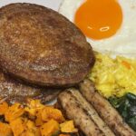 Close-up of a rustic breakfast spread with crispy golden oat pancakes, roasted sweet potatoes, browned chicken sausages, sautéed spinach, turmeric rice, and a runny fried egg.