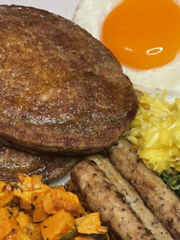 Close-up of a rustic breakfast spread with crispy golden oat pancakes, roasted sweet potatoes, browned chicken sausages, sautéed spinach, turmeric rice, and a runny fried egg.