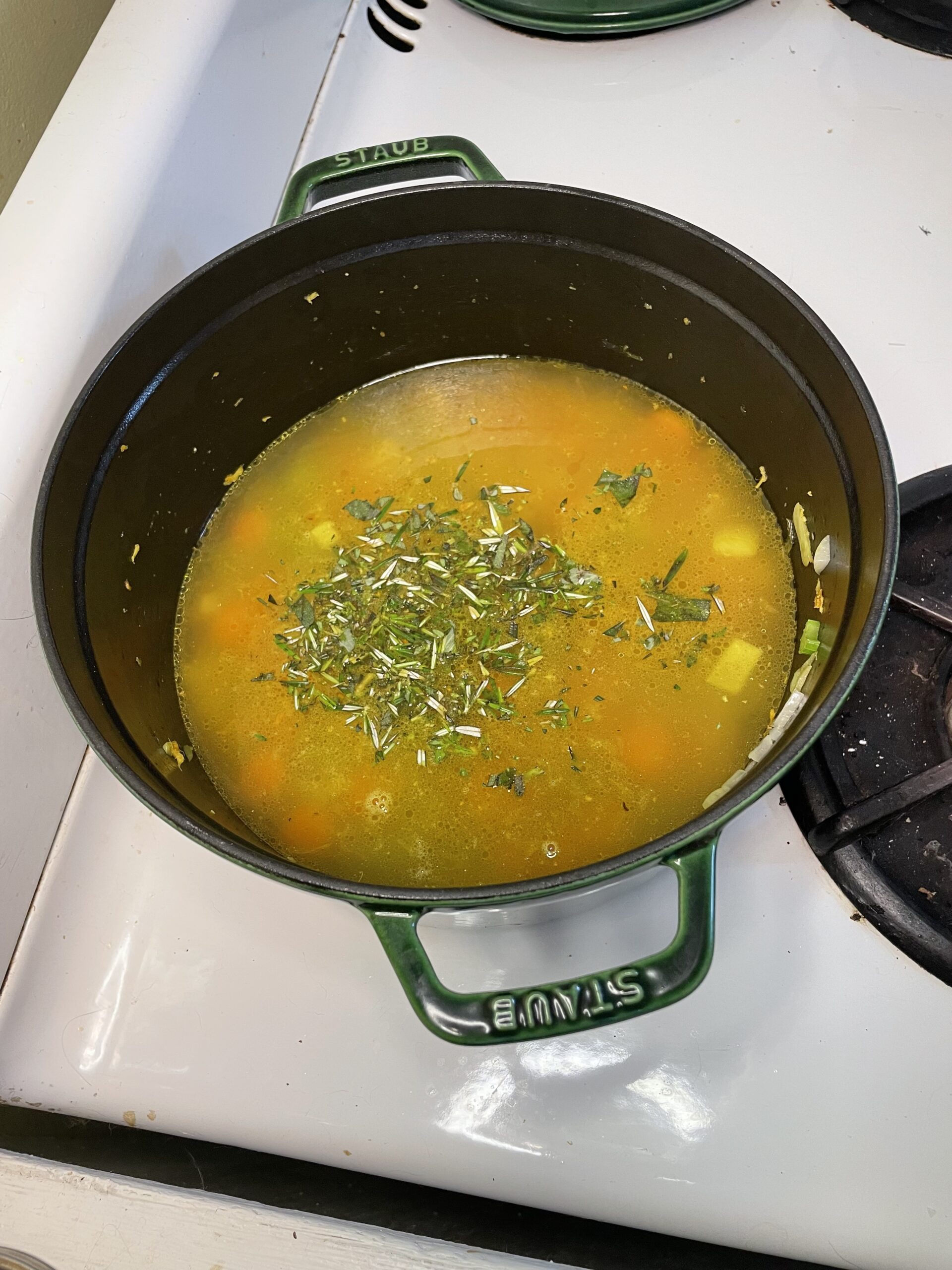 Overhead view of a green Staub pot filled with a vibrant broth-based soup, topped generously with fresh herbs.
