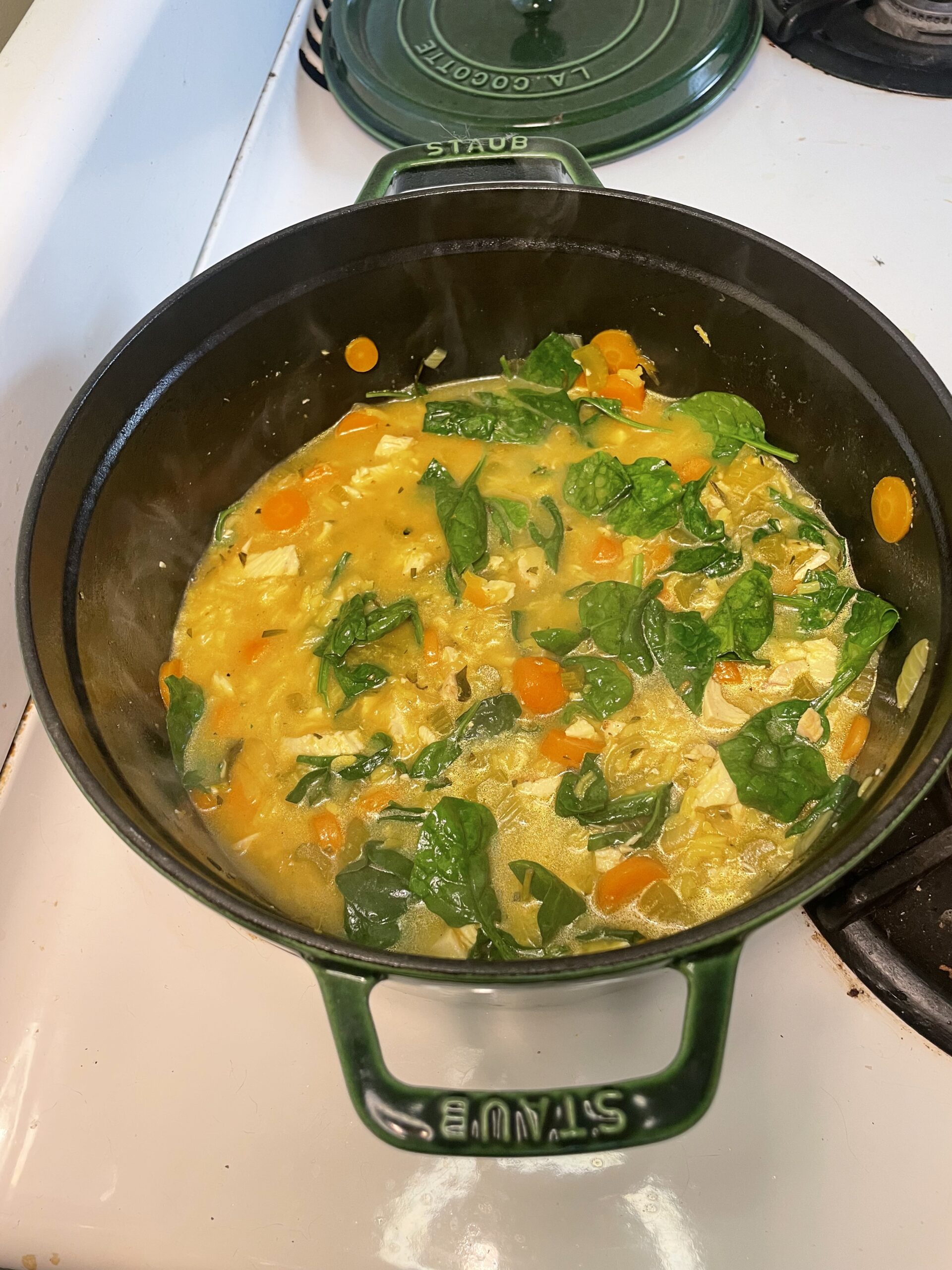 Cooking process of hearty soup in a green Staub pot, now thickened with colorful vegetables and bright green spinach leaves.