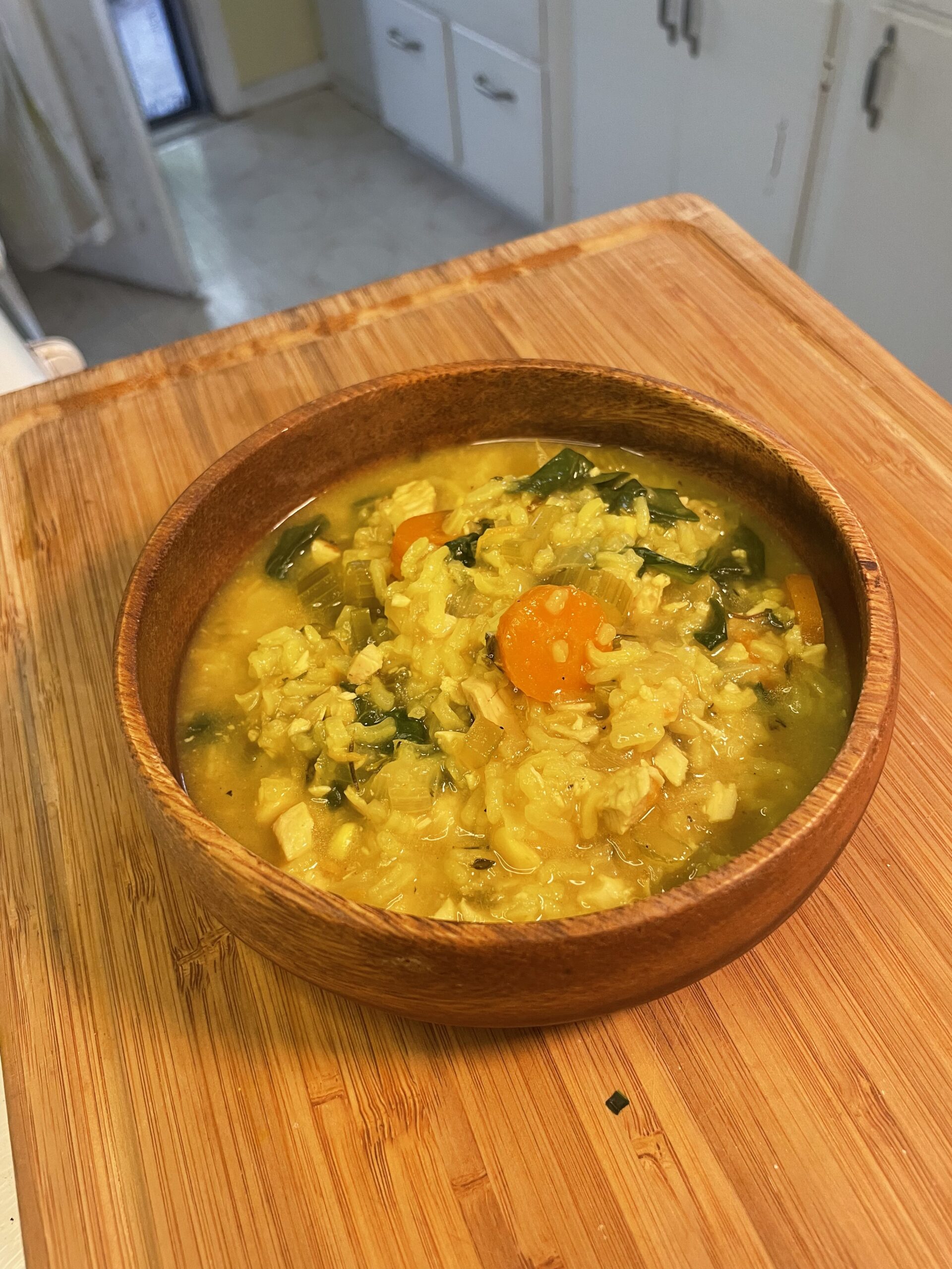 A rustic wooden bowl filled with golden, nourishing soup featuring chunks of carrots, greens, and hearty grains placed on a wooden countertop.