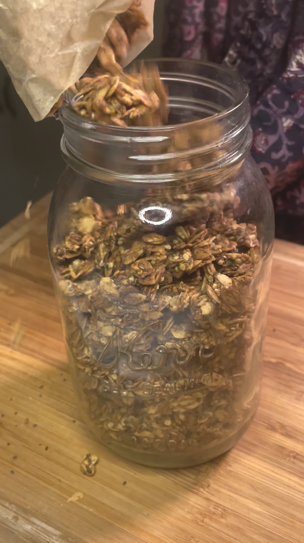 Pouring crunchy maple oat and seed granola into a glass jar for storage.