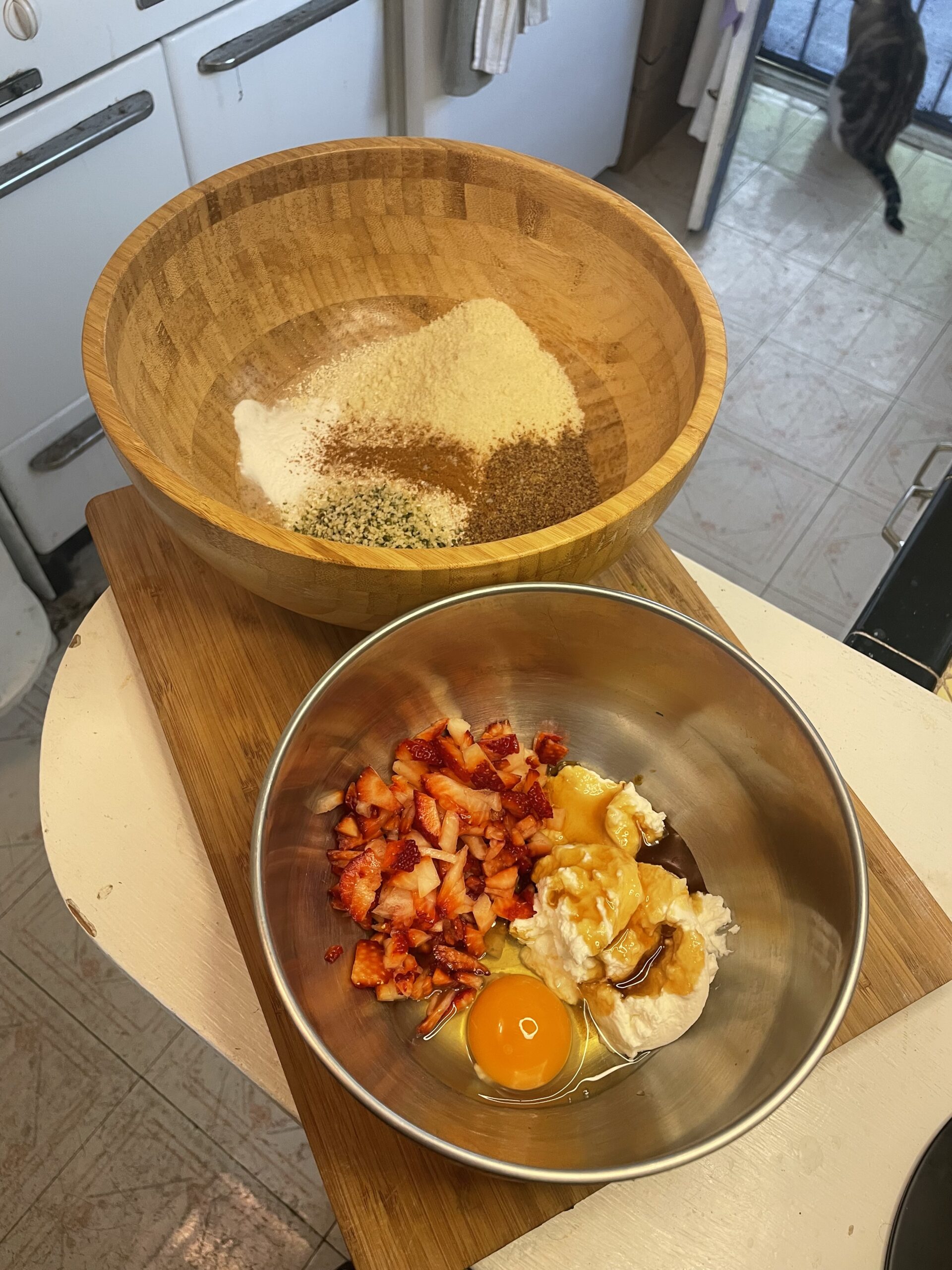 Two mixing bowls—one filled with flours and grains, the other with chopped strawberries, egg, and yogurt—sitting side by side on a cutting board.