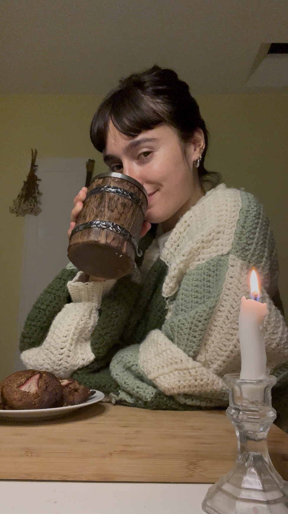 A woman in a chunky green and white sweater smiles while sipping from a rustic mug, with muffins and a lit candle on the table in front of her.