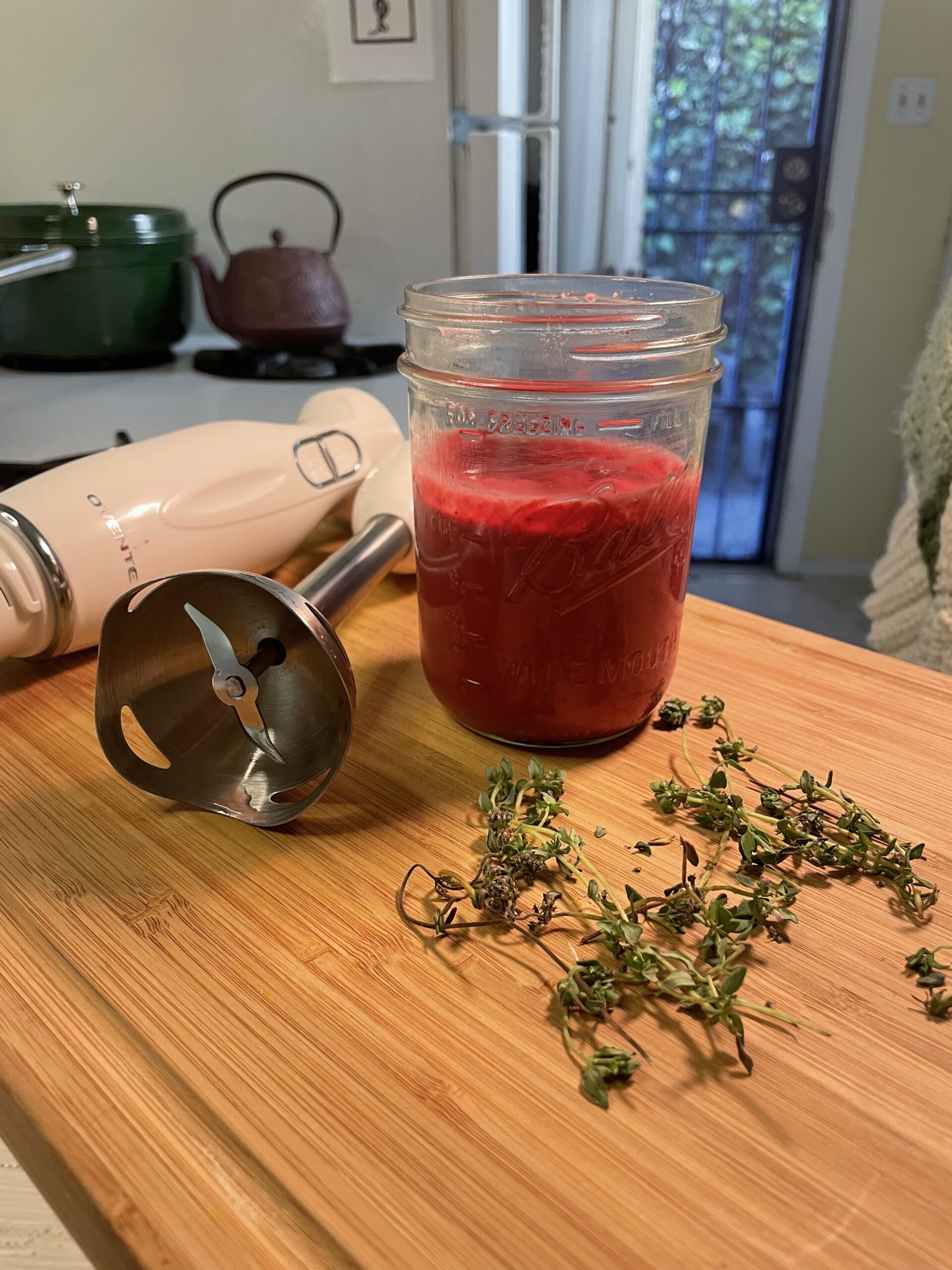 A glass mason jar filled with bright pink sauce next to a white immersion blender and scattered fresh herbs on a wooden cutting board.