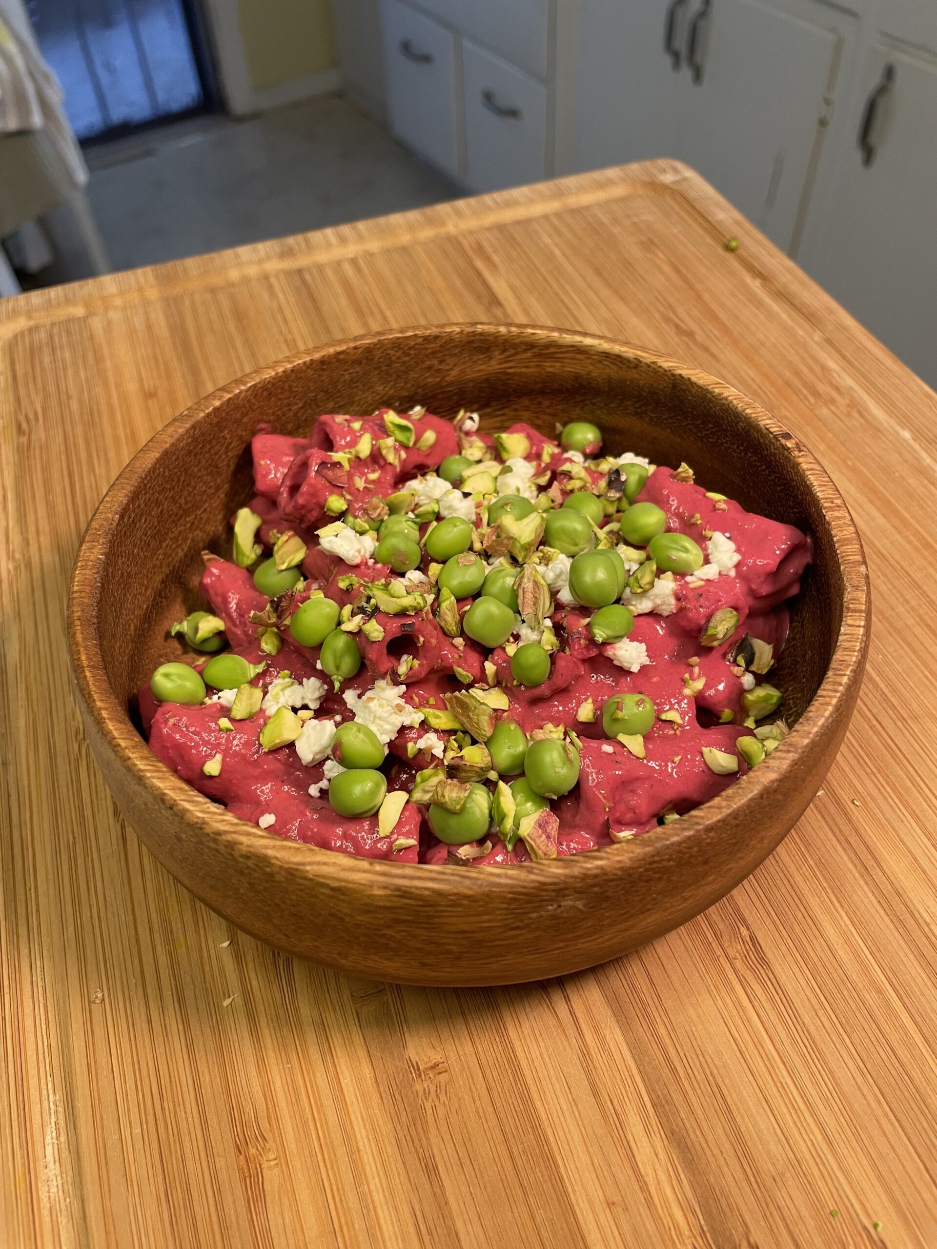 Pink pasta dish served in a wooden bowl, generously topped with fresh peas, crumbled cheese, and chopped pistachios, placed on a wooden countertop.