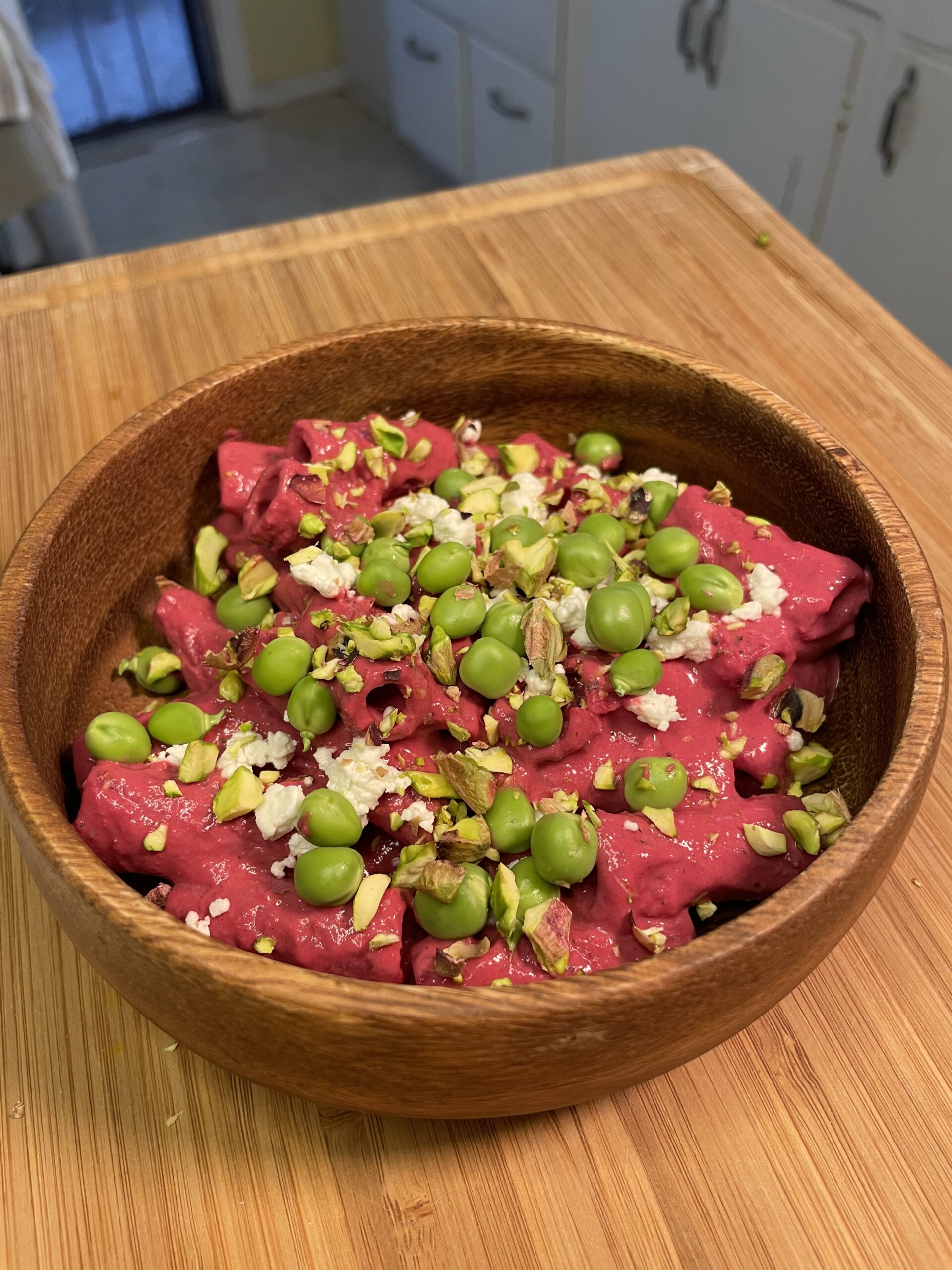 Close-up of vibrant pink pasta garnished with fresh green peas, crumbled cheese, and crushed pistachios in a rustic wooden bowl.