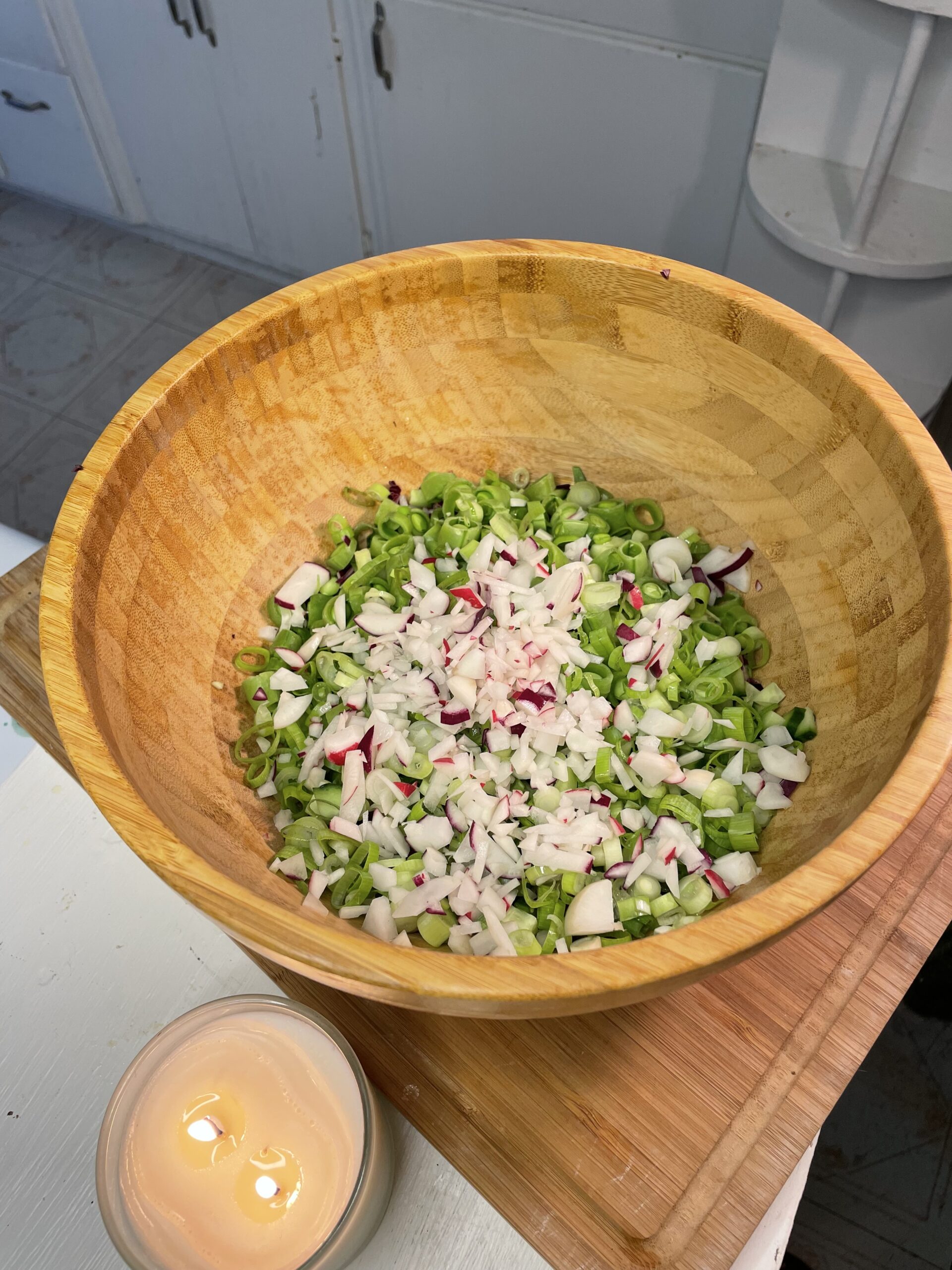 Freshly chopped veggies in a large wooden bowl beside a lit candle, setting a cozy kitchen atmosphere for homemade slaw