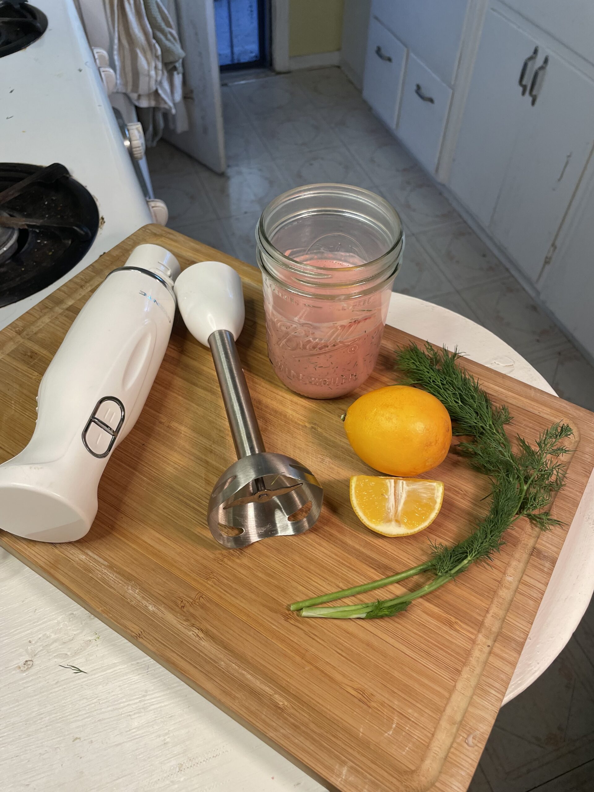 Ingredients for homemade dressing on a cutting board with immersion blender, lemon, fresh dill, and a pink sauce in a mason jar
