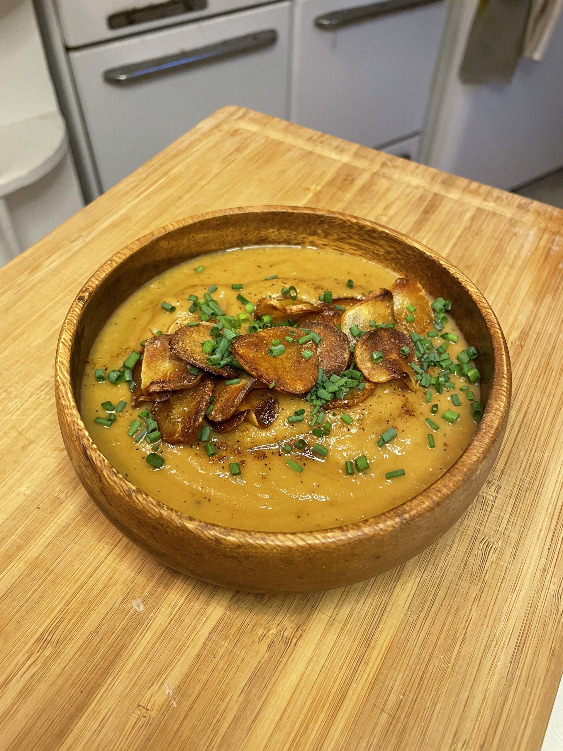 Finished creamy roasted parsnip soup served in a wooden bowl, topped with golden-brown parsnip chips, fresh chives, and a sprinkle of spices.