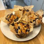 These are the finished baked goods—five individual blueberry lavender coffee cakes placed on a white plate. They're wrapped in rustic parchment muffin liners and topped with a golden, crumbly streusel topping that's caramelized in places. The vibrant blueberries have bubbled up through the top, creating a beautiful contrast against the golden brown topping.