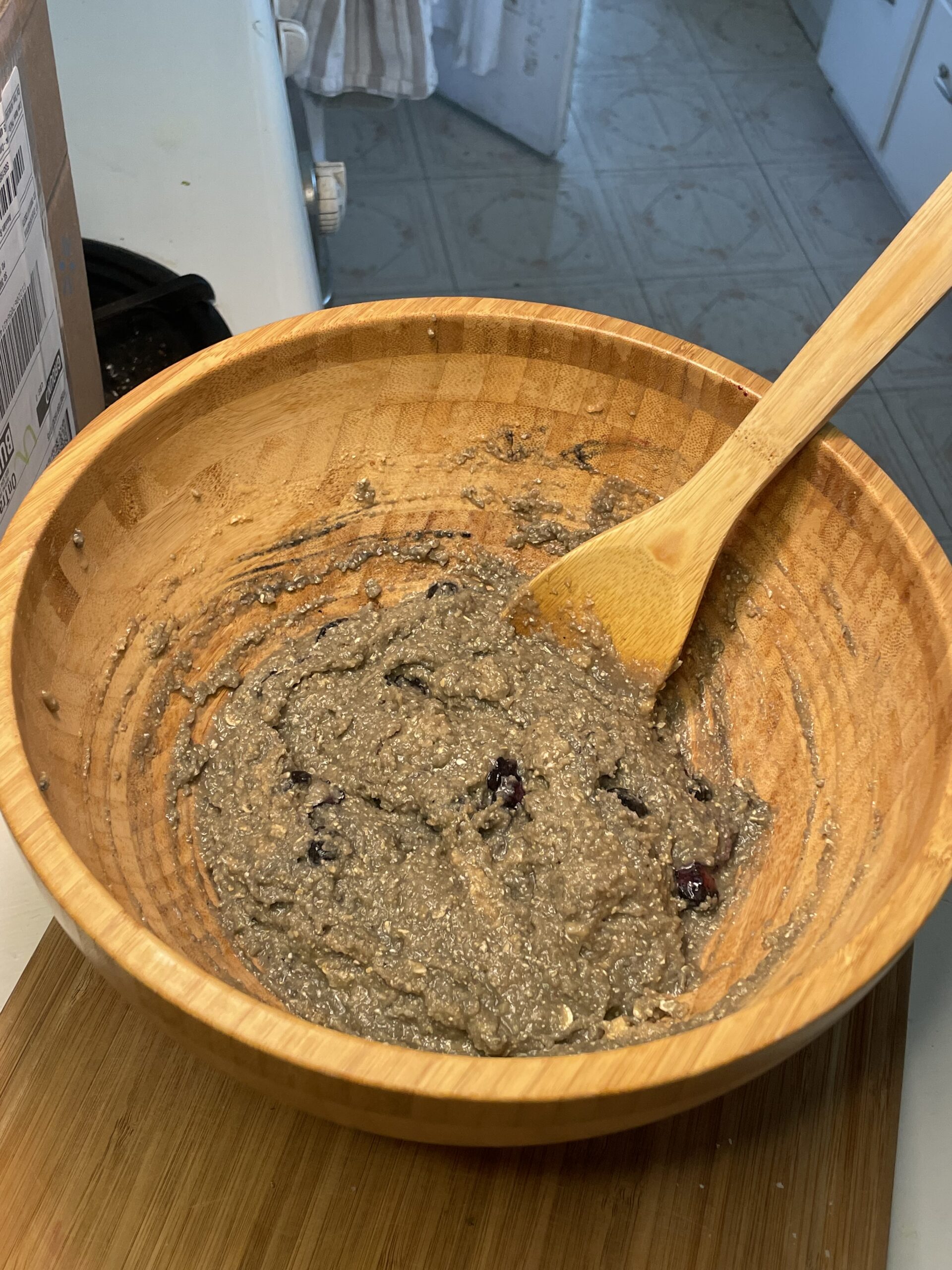 This is a wooden mixing bowl containing a thick, dark batter. The batter appears to include oats or almond meal, visible chia or flax seeds, and dark-colored berries—likely blueberries. The consistency looks moist and slightly chunky. A wooden spoon rests in the bowl, indicating the batter is in the preparation or mixing stage.