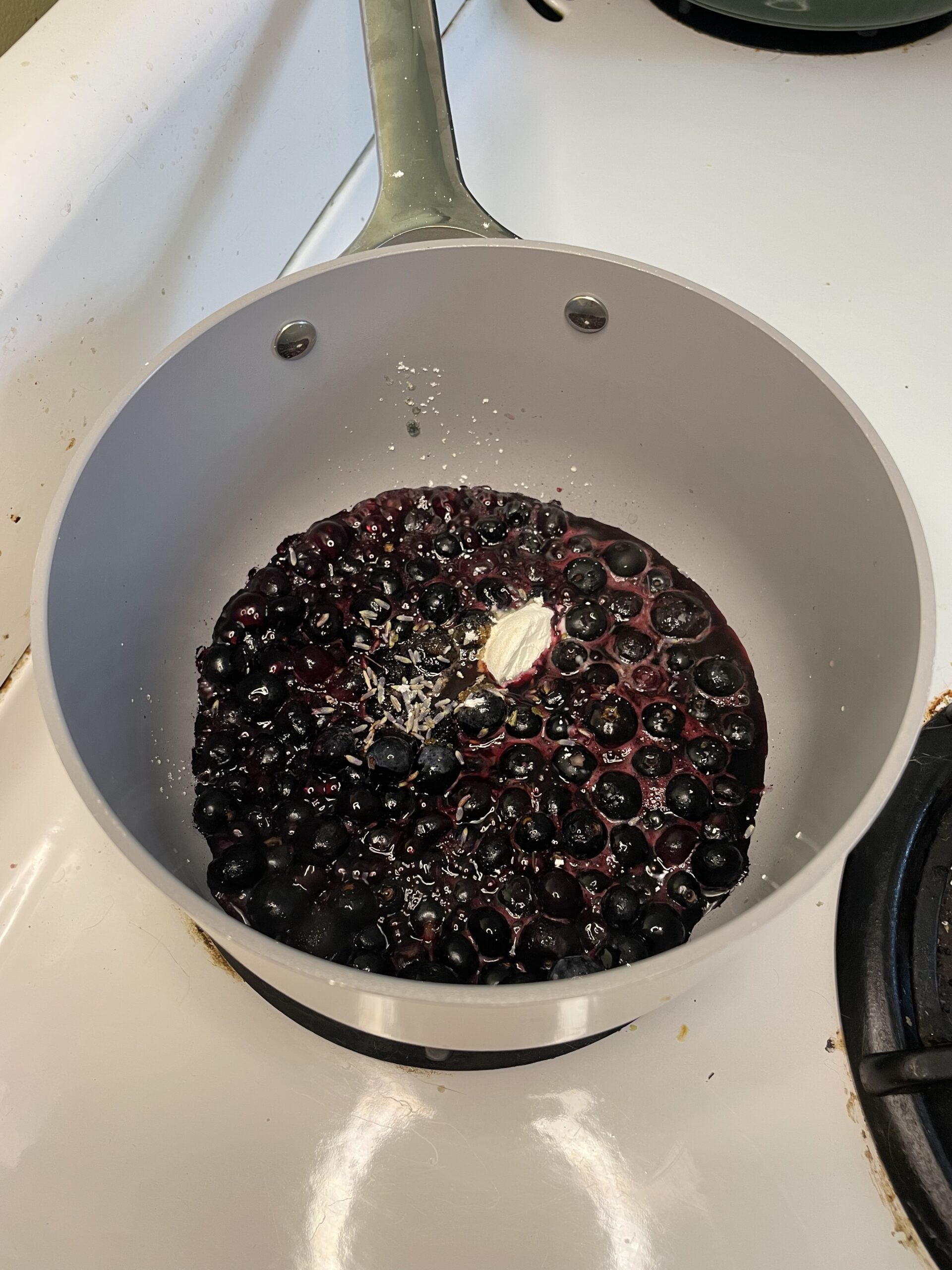 This image shows the process of making a blueberry-lavender compote or filling on the stovetop. In a light-colored saucepan, fresh or frozen blueberries are being cooked down. The berries are starting to burst and release their juices, forming a rich, deep-purple mixture.