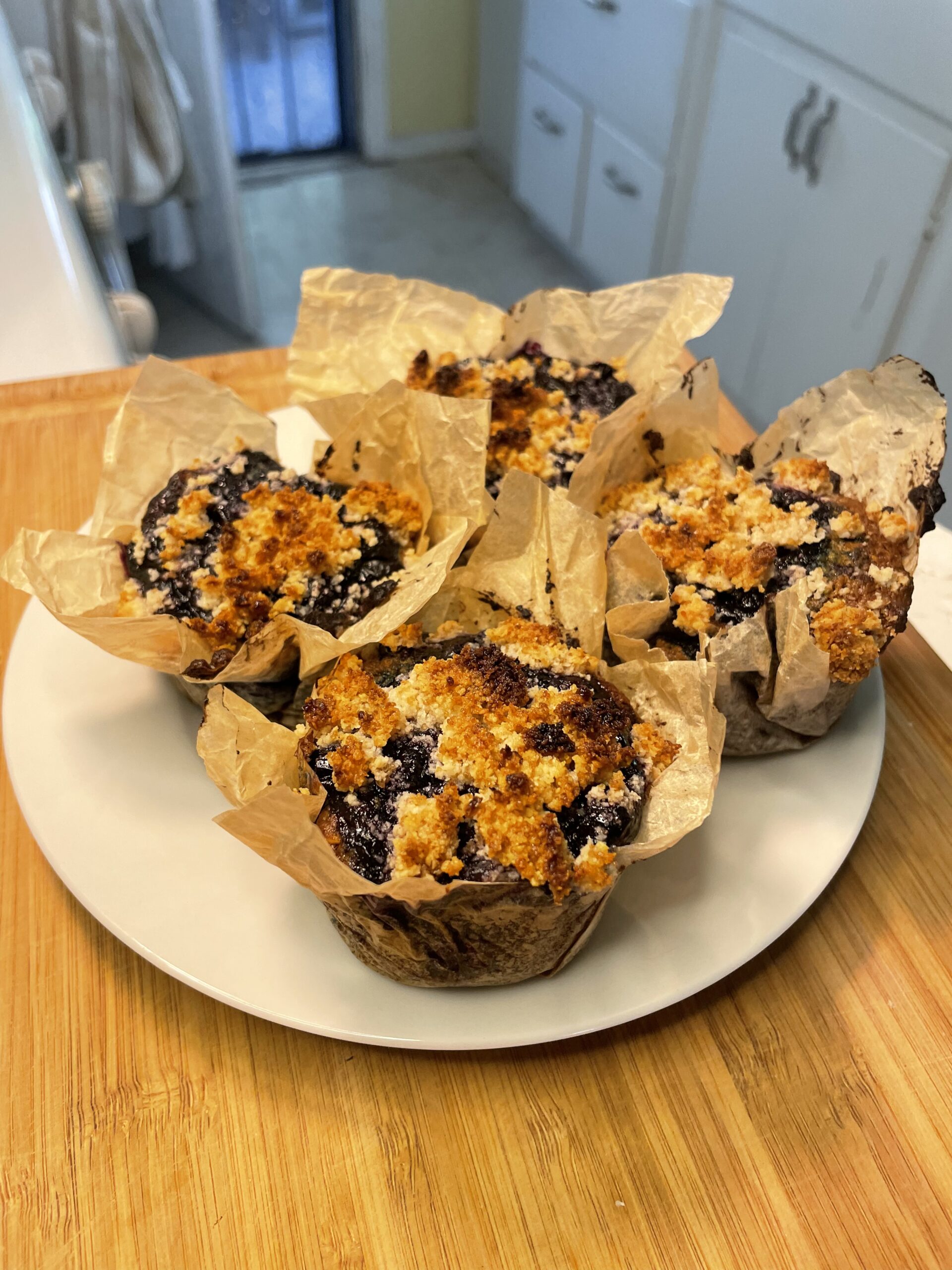 These are the finished baked goods—four individual blueberry lavender coffee cakes placed on a white plate. They're wrapped in rustic parchment muffin liners and topped with a golden, crumbly streusel topping that's caramelized in places. The vibrant blueberries have bubbled up through the top, creating a beautiful contrast against the golden brown topping.