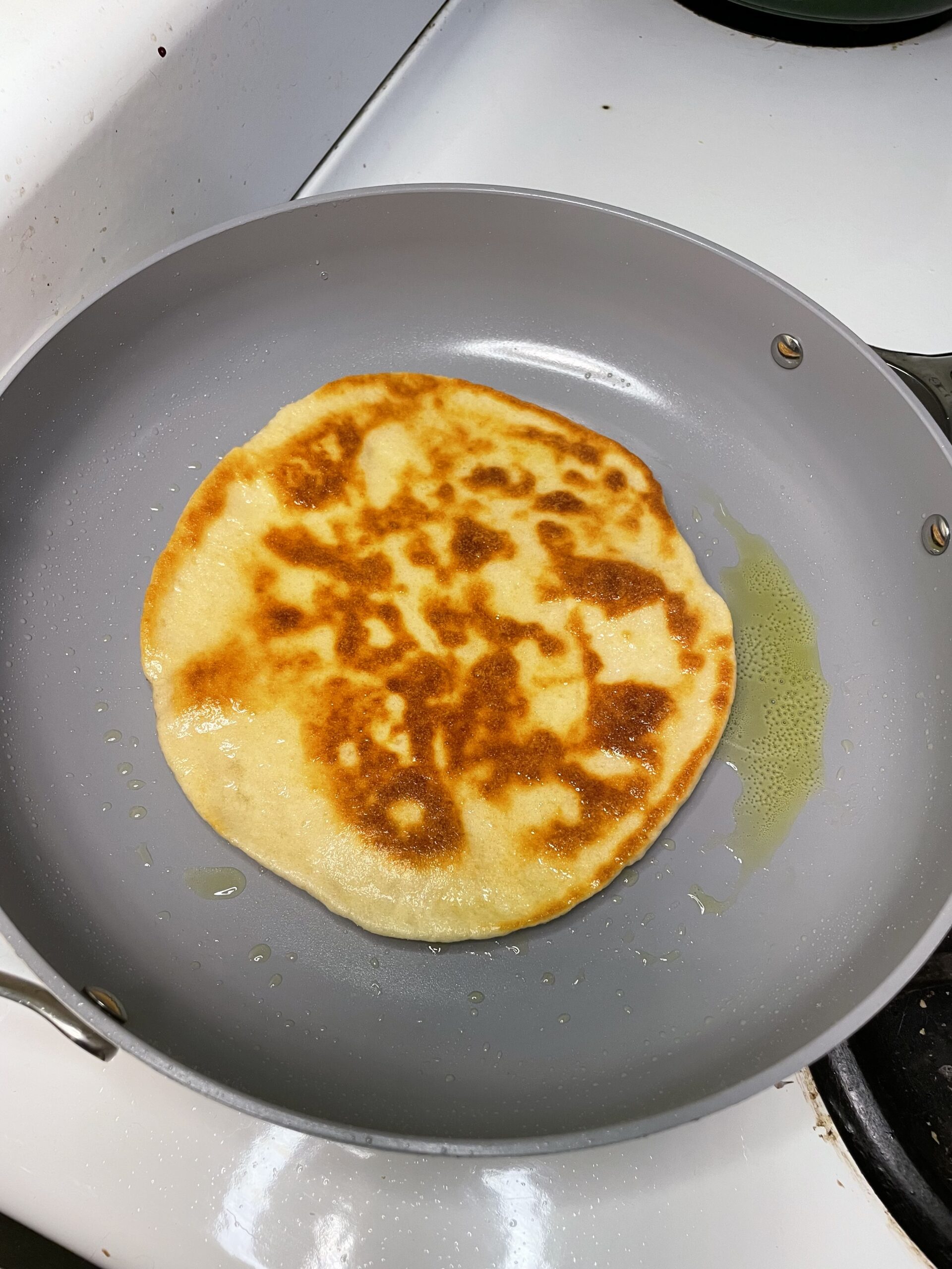 Flatbread rests in a non-stick grey cooking pan filled with olive oil, a top a white stovetop.  This is the first step in creating the Enchanted Rainbow Veggie Flatbread.
