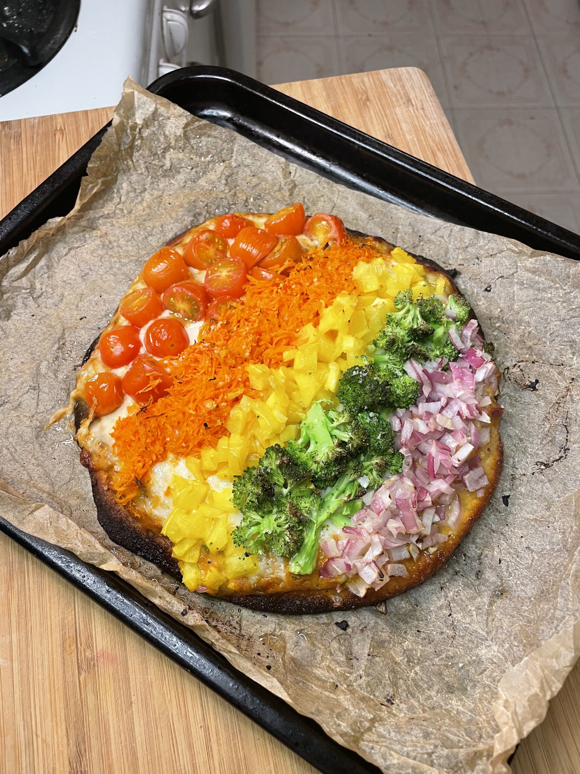 This image features a beautifully vibrant Enchanted Rainbow Veggie Flatbread fresh from the oven. Baked on parchment paper atop a dark baking tray, the flatbread is visually striking with a rainbow-like arrangement of colorful vegetables, layered in neat rows: Red cherry tomatoes (halved) form the top row, blistered and juicy from roasting,
shredded orange carrots come next, slightly softened and bright, yellow bell peppers, diced, add a sunny burst of color, roasted green broccoli florets provide a hearty, earthy contrast, and finely chopped red onions complete the rainbow, adding a touch of sweetness and crunch.
The flatbread base has a golden, slightly crisped edge with a bit of charring on the bottom, giving it a rustic, wood-fired appearance. Melted cheese peeks out beneath the veggie layers, binding everything together. The presentation is playful, nutritious, and perfect for a whimsical, garden-to-table meal.