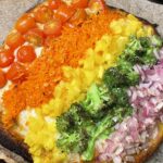 This image features a beautifully vibrant Enchanted Rainbow Veggie Flatbread fresh from the oven. Baked on parchment paper atop a dark baking tray, the flatbread is visually striking with a rainbow-like arrangement of colorful vegetables, layered in neat rows: Red cherry tomatoes (halved) form the top row, blistered and juicy from roasting, shredded orange carrots come next, slightly softened and bright, yellow bell peppers, diced, add a sunny burst of color, roasted green broccoli florets provide a hearty, earthy contrast, and finely chopped red onions complete the rainbow, adding a touch of sweetness and crunch. The flatbread base has a golden, slightly crisped edge with a bit of charring on the bottom, giving it a rustic, wood-fired appearance. Melted cheese peeks out beneath the veggie layers, binding everything together. The presentation is playful, nutritious, and perfect for a whimsical, garden-to-table meal.