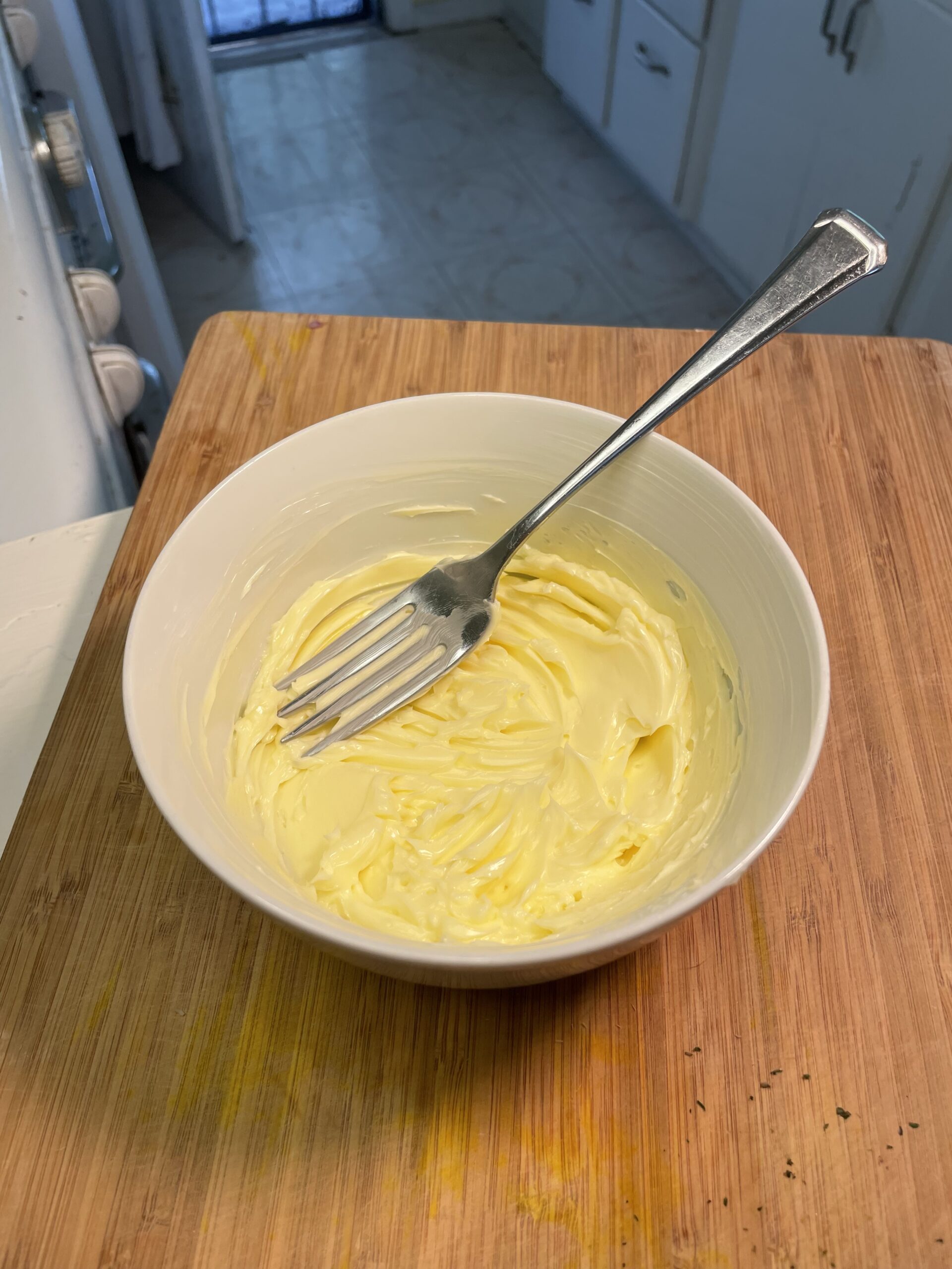 This image shows a white bowl, filled with whipped, softened butter, set on a wooden cutting board.  A silver spoon rests in the bowl to demonstrate the tool used to whip the butter into its texture.